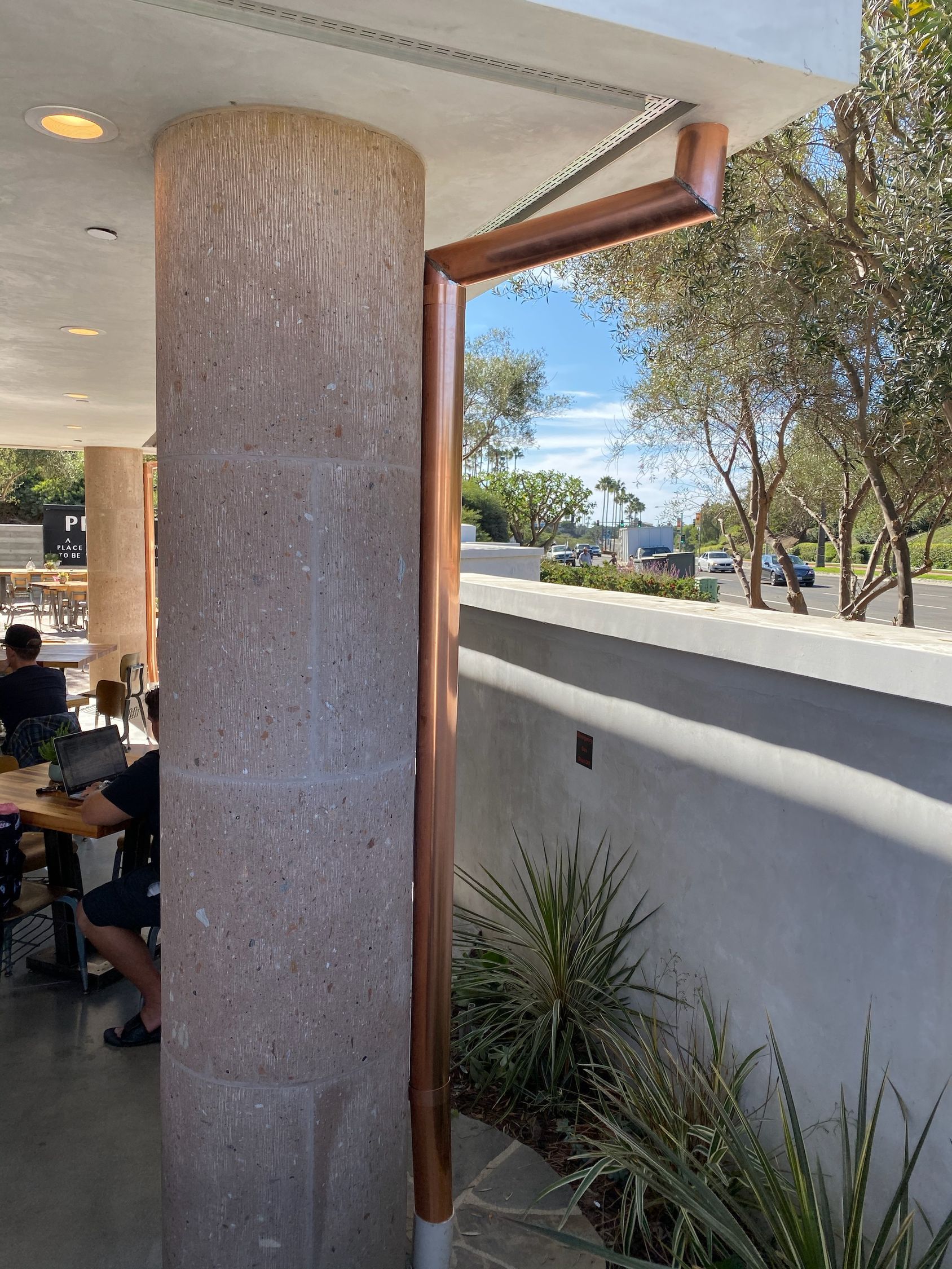 Copper downspout on a stone column. Exterior view with plants and people dining in the background.