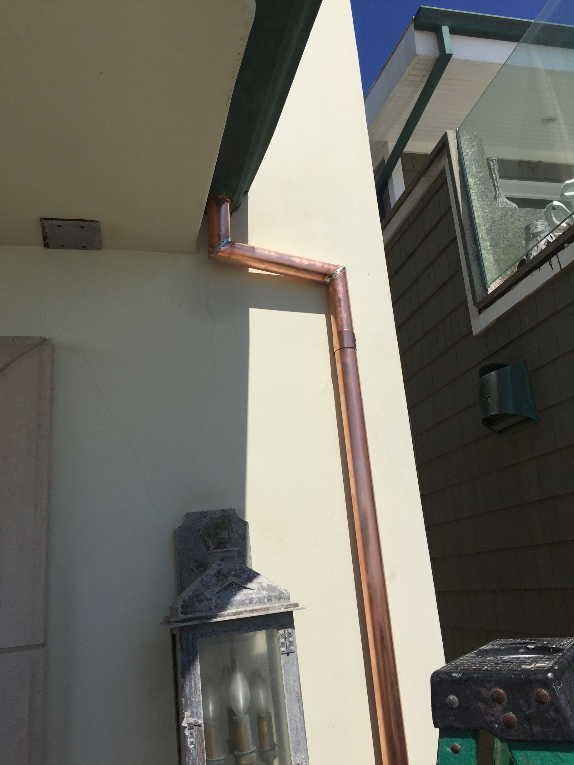 Copper rain gutter on a light-colored building exterior. A lantern is below. Bright sunlight.
