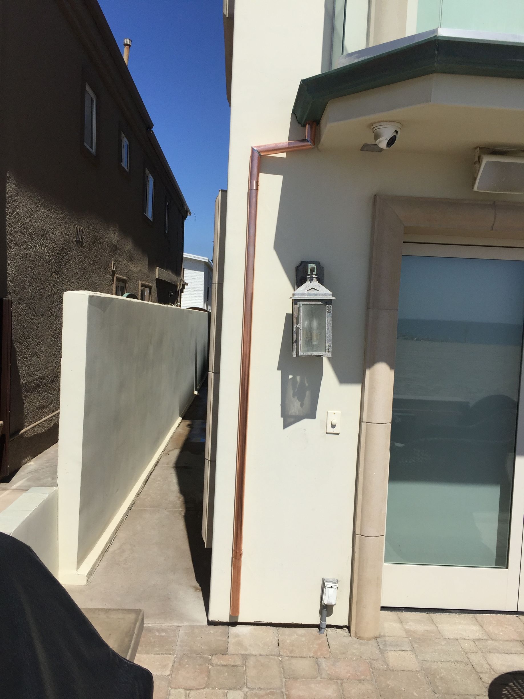 Narrow alley between two buildings, one beige and one brown. A copper pipe runs up the beige building's exterior.