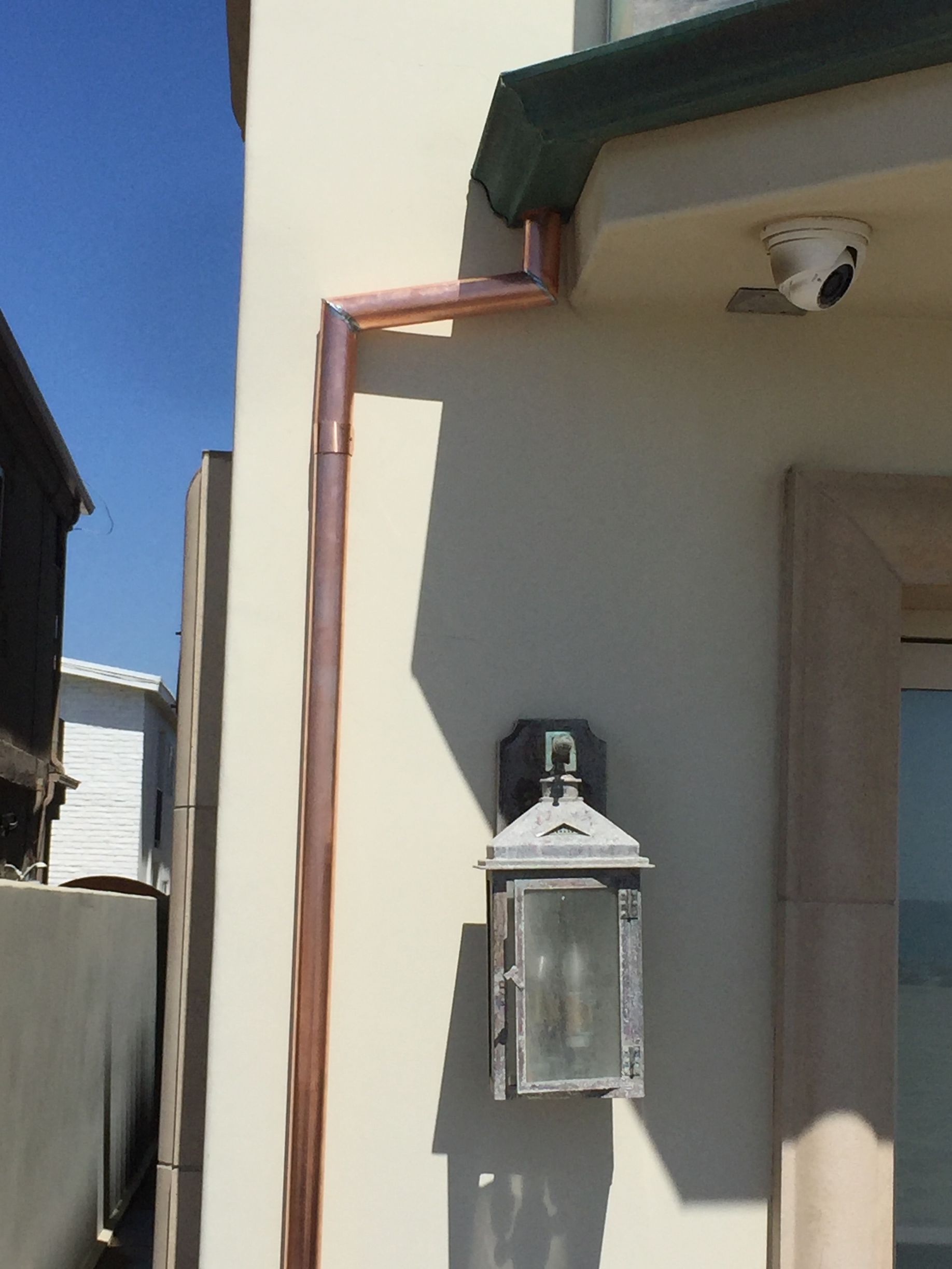 Copper gutter and downspout on a beige building next to a lamp. Security camera above.