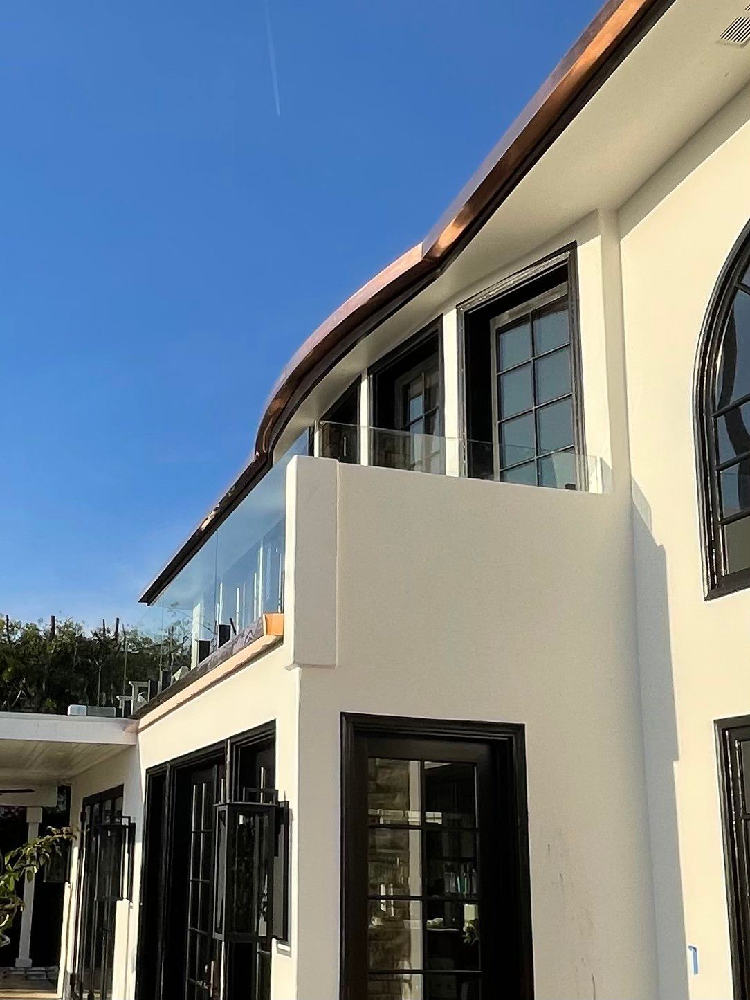 White building with black trim and curved roof, balcony with glass railing, against blue sky.