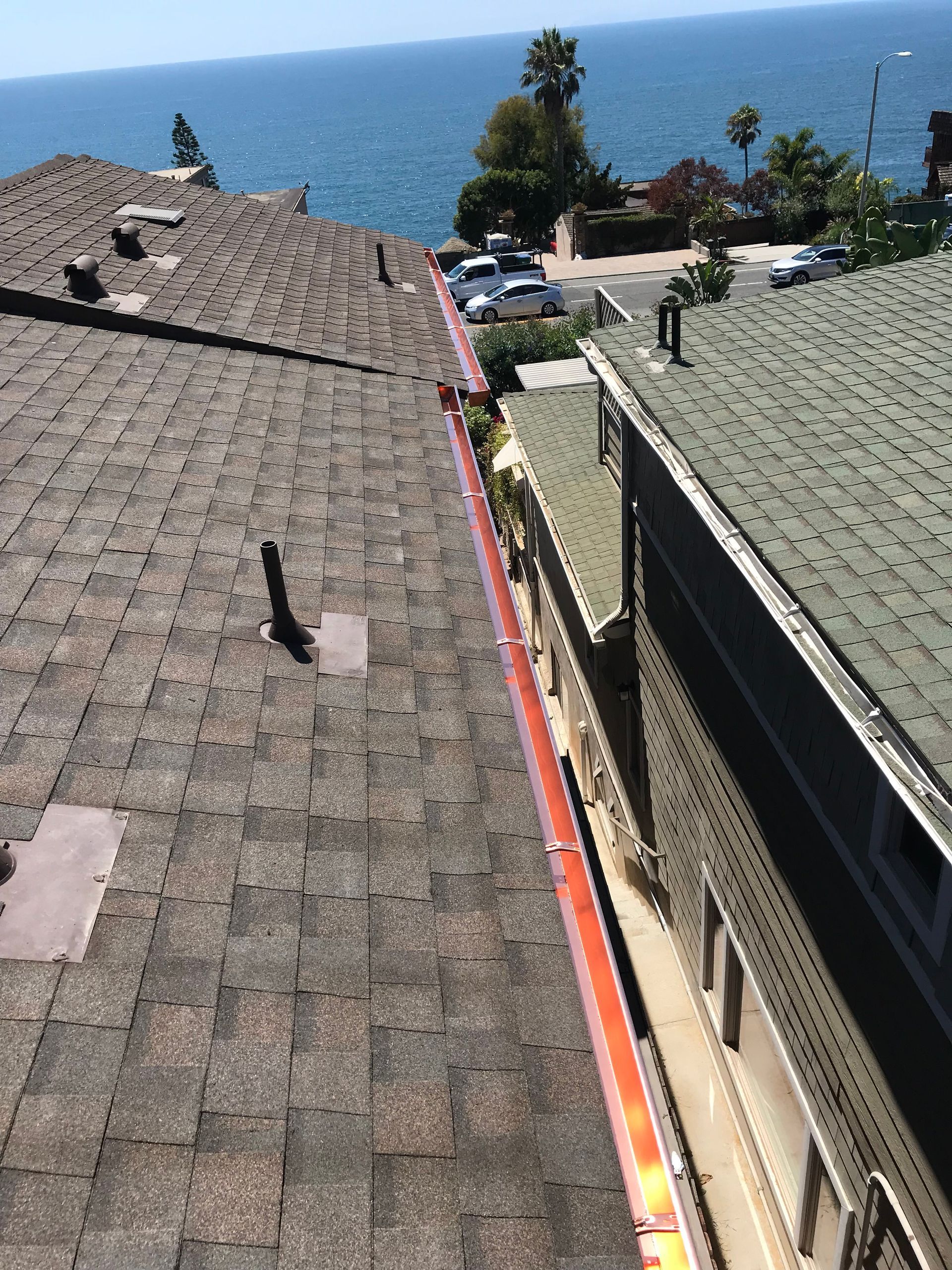 Rooftop view overlooking the ocean and street. Asphalt shingles, red gutters, blue water, white cars, and trees.