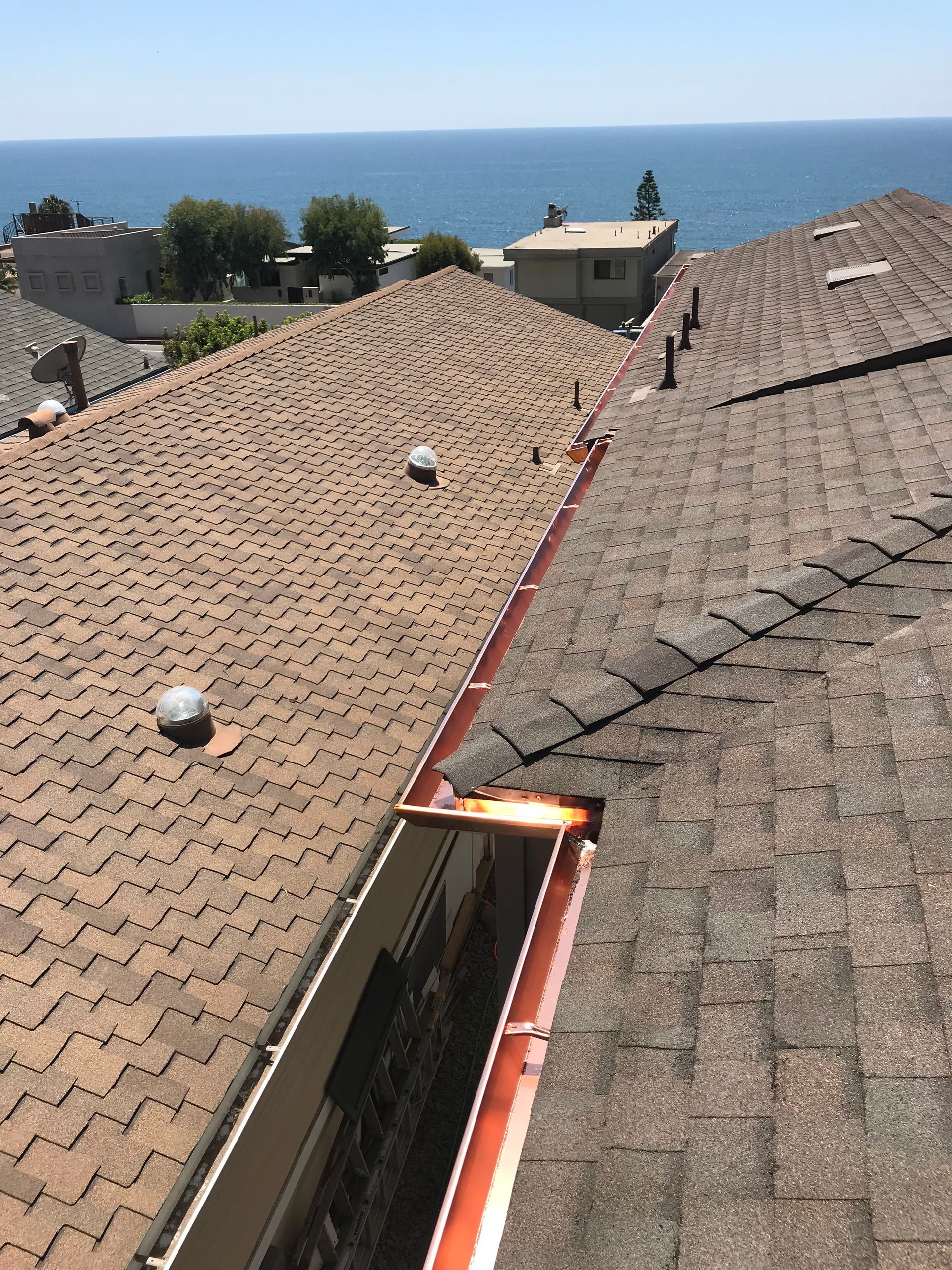 View of a brown shingled roof with a copper gutter, ocean visible in the distance under a bright blue sky.