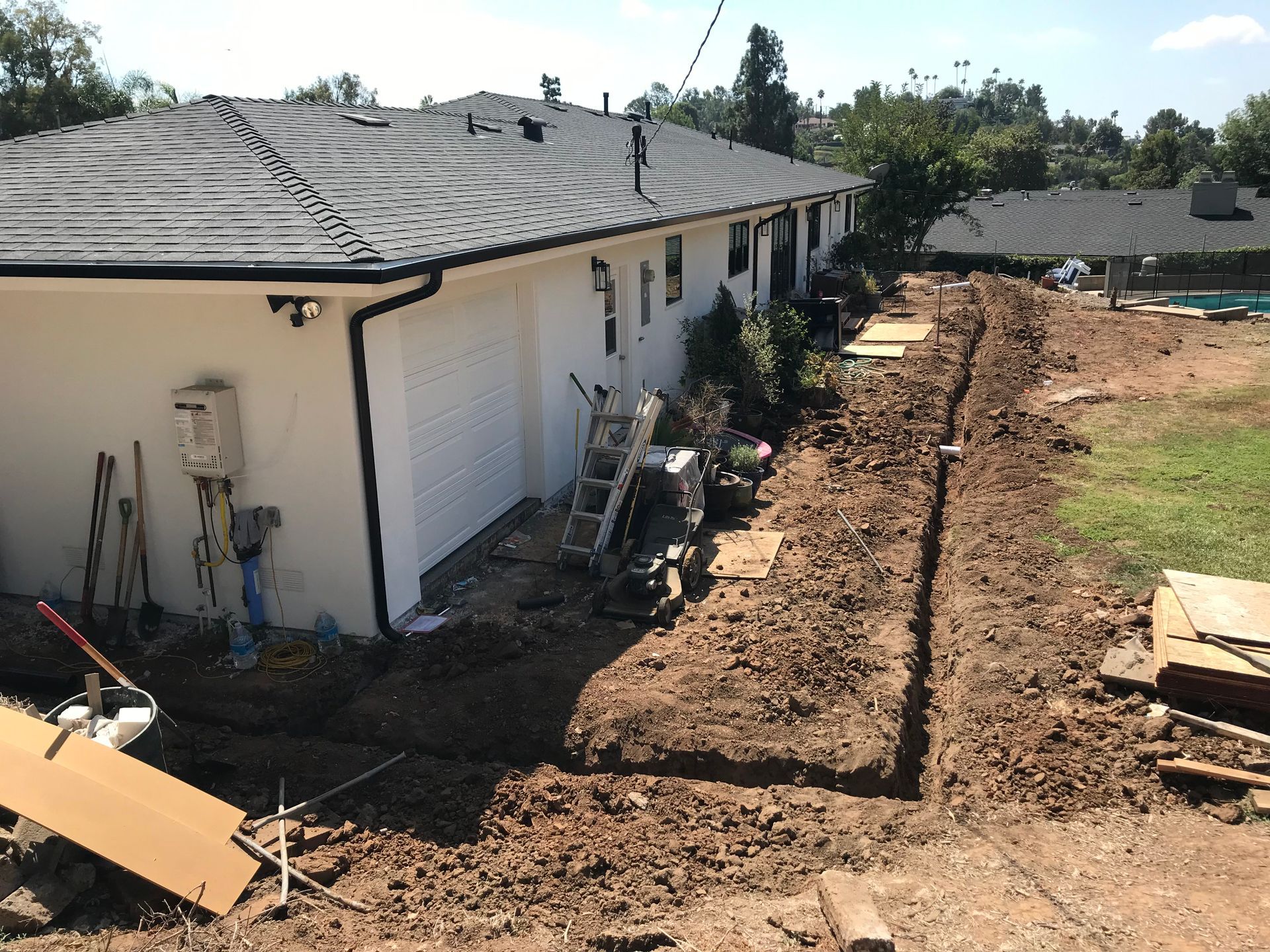 Trench dug along a white house, construction materials and tools nearby on a sunny day.