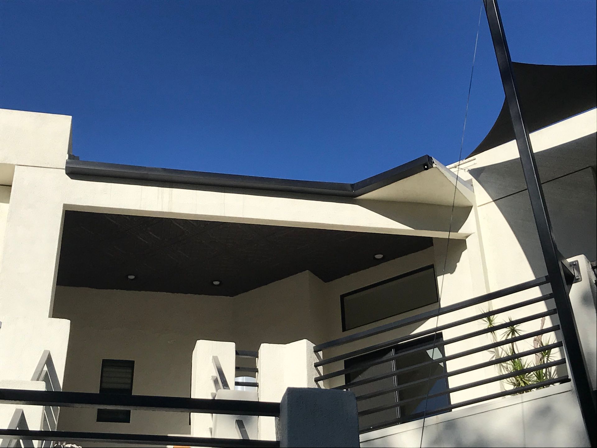Modern white house with black roof, dark wood ceiling, and black railing against a bright blue sky.