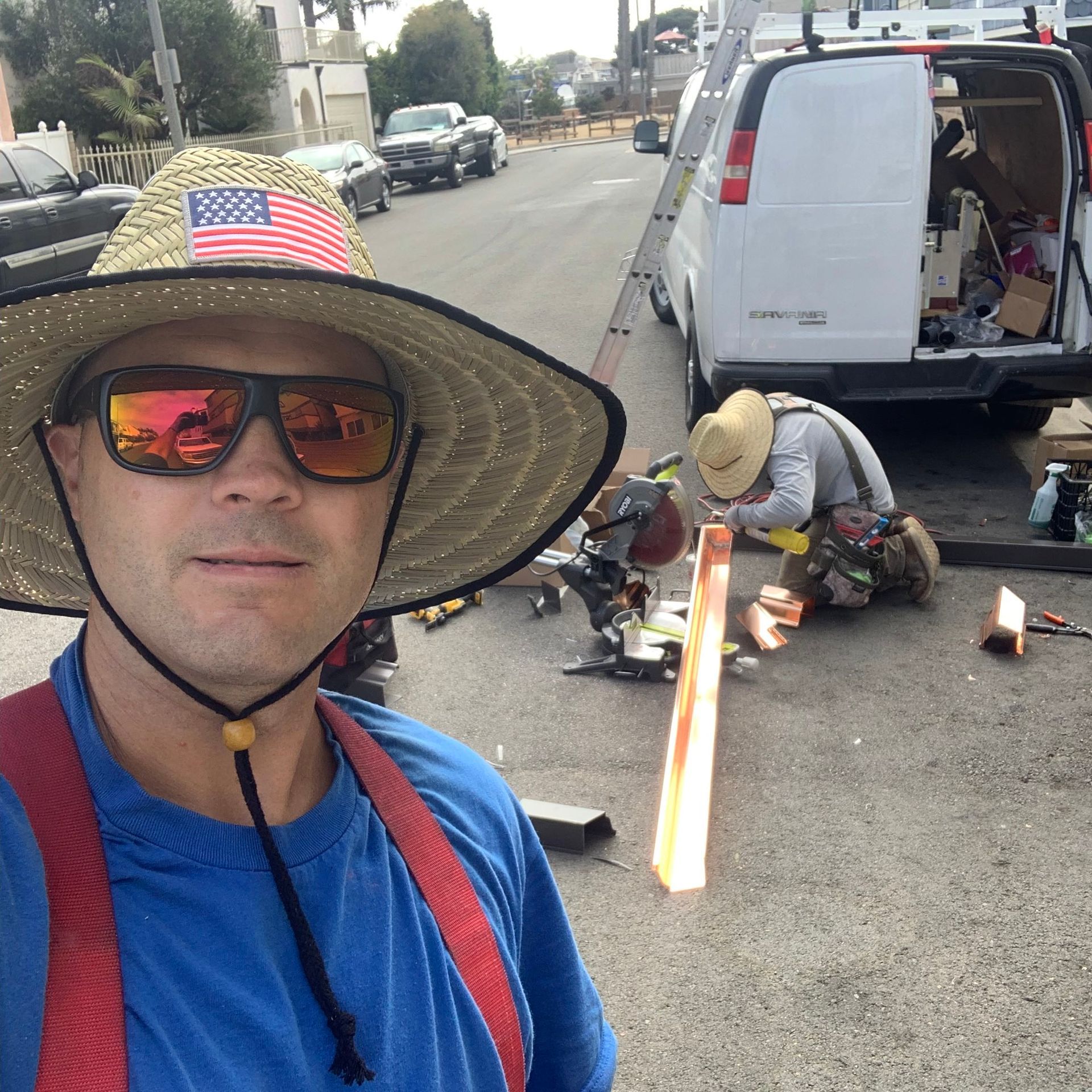 Man in hat with sunglasses takes selfie. Two other workers repair equipment near a white van on a city street.