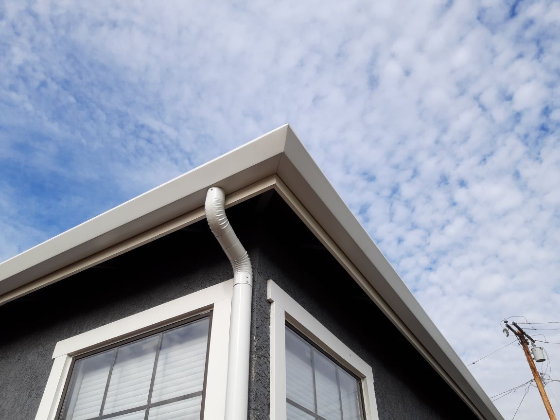 Corner of a dark building with white trim and a gutter against a cloudy blue sky.