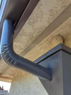 Dark gray downspout bends from brown gutter on a beige stucco wall.