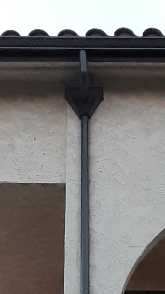 Dark grey rain gutter system on a light stucco wall, with a downward-facing triangular component.