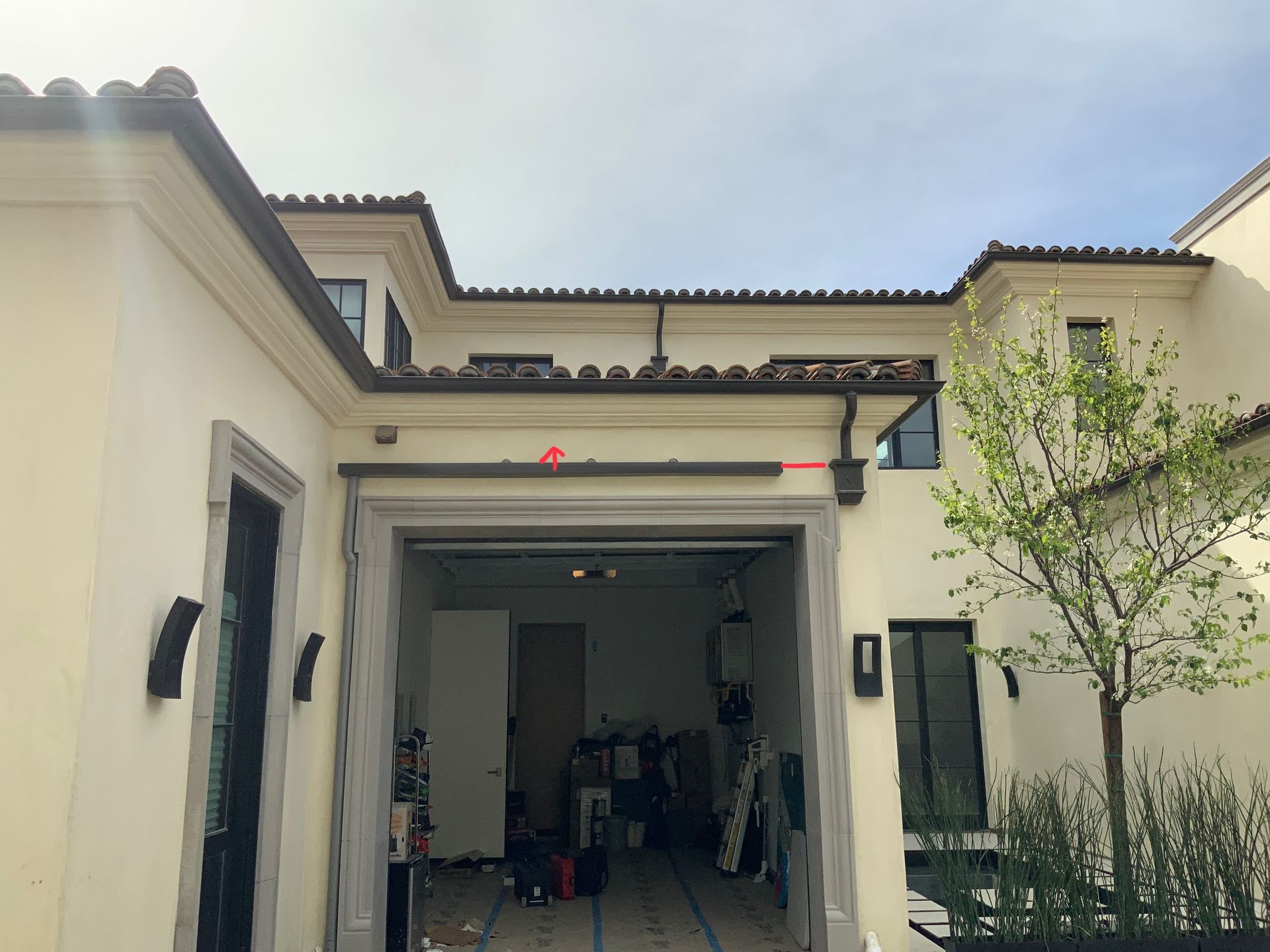 Beige house exterior with an open garage, brown roof, and small tree.