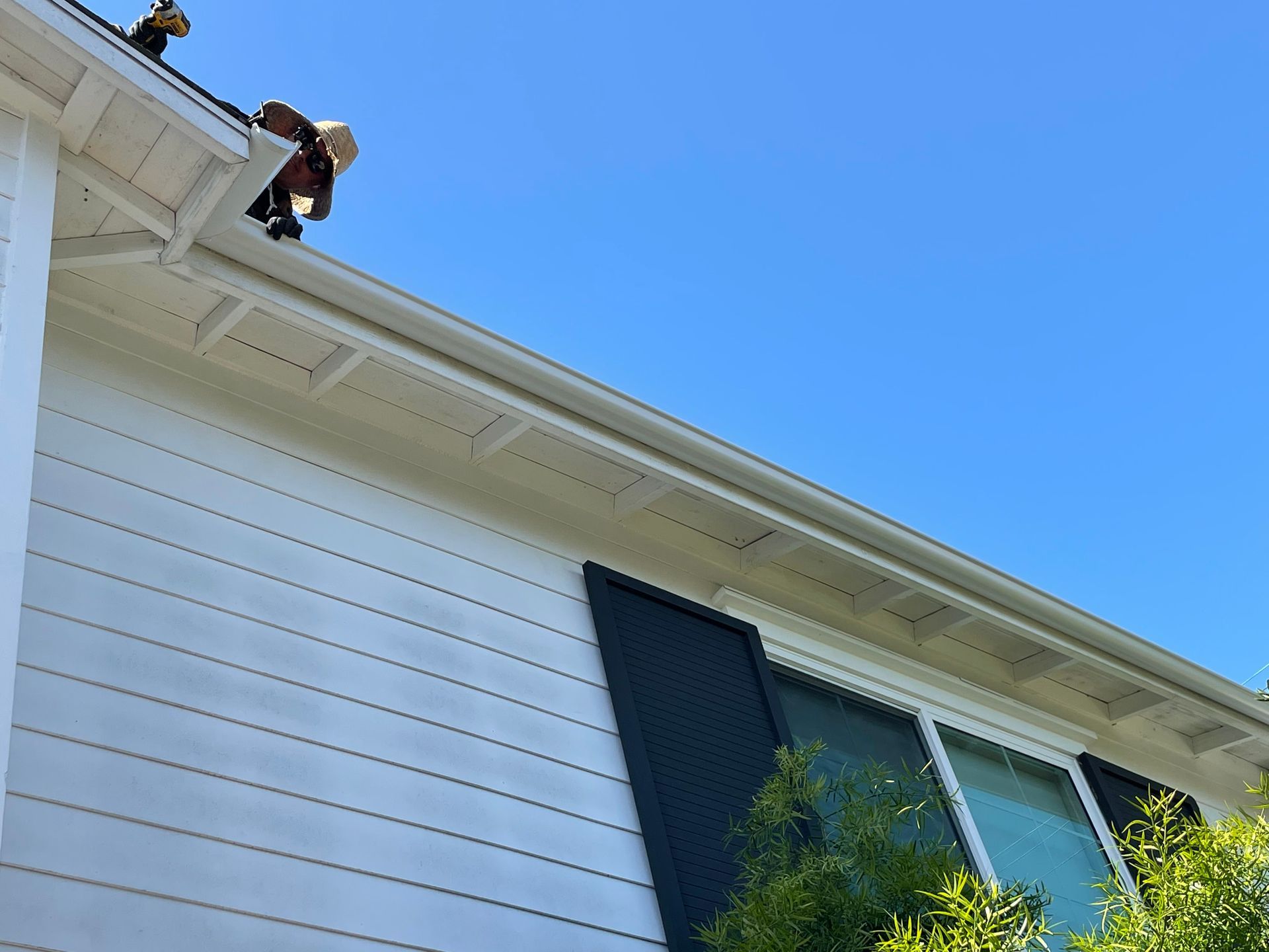Person cleaning a white gutter on a house, blue sky.