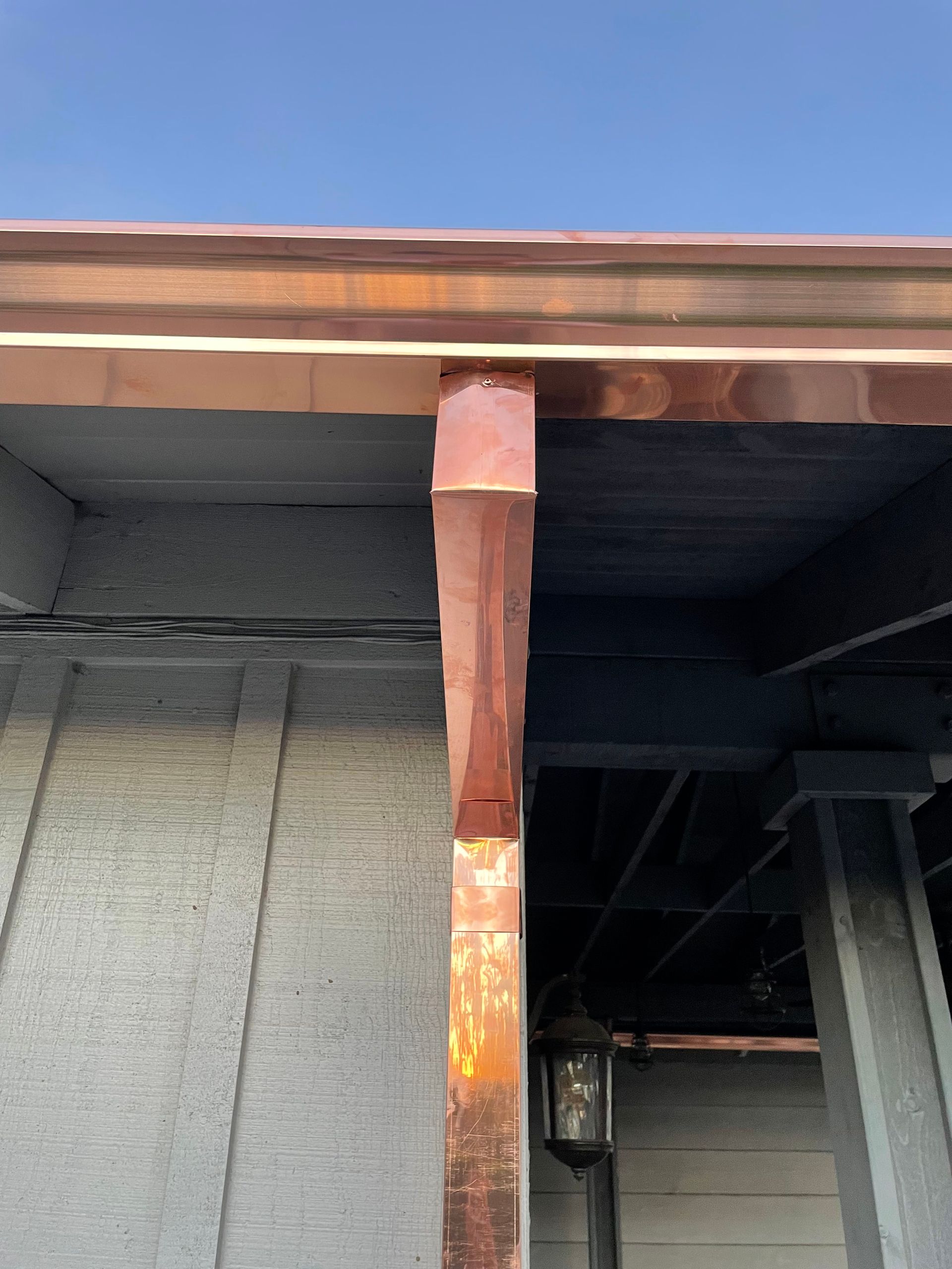 Copper-clad column supports a copper gutter on a building exterior; light gray siding and dark ceiling.