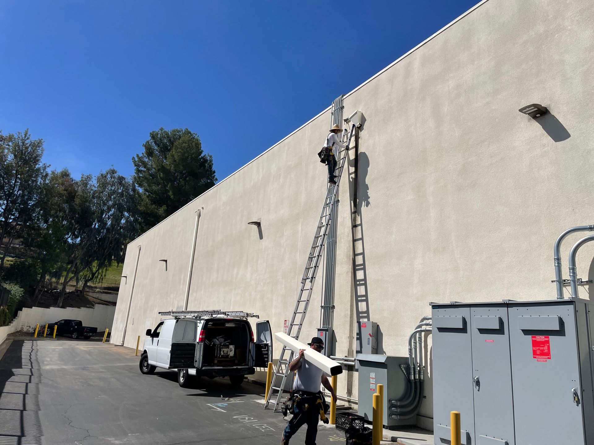 Two workers on ladders near a building wall; one near roof. A van and conduit pipe are at the ground level.