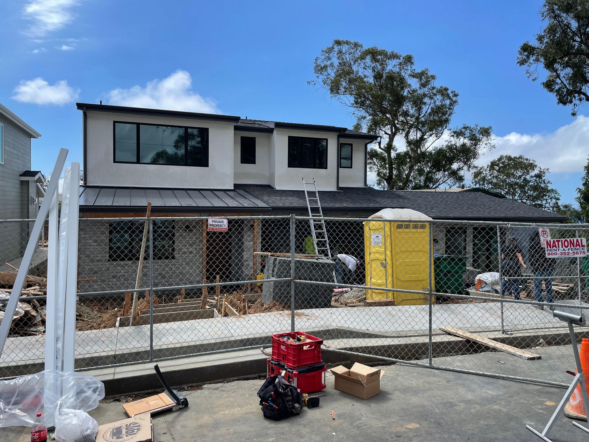 Construction site with two-story building. Fenced area, portable toilet, tools, and blue sky.