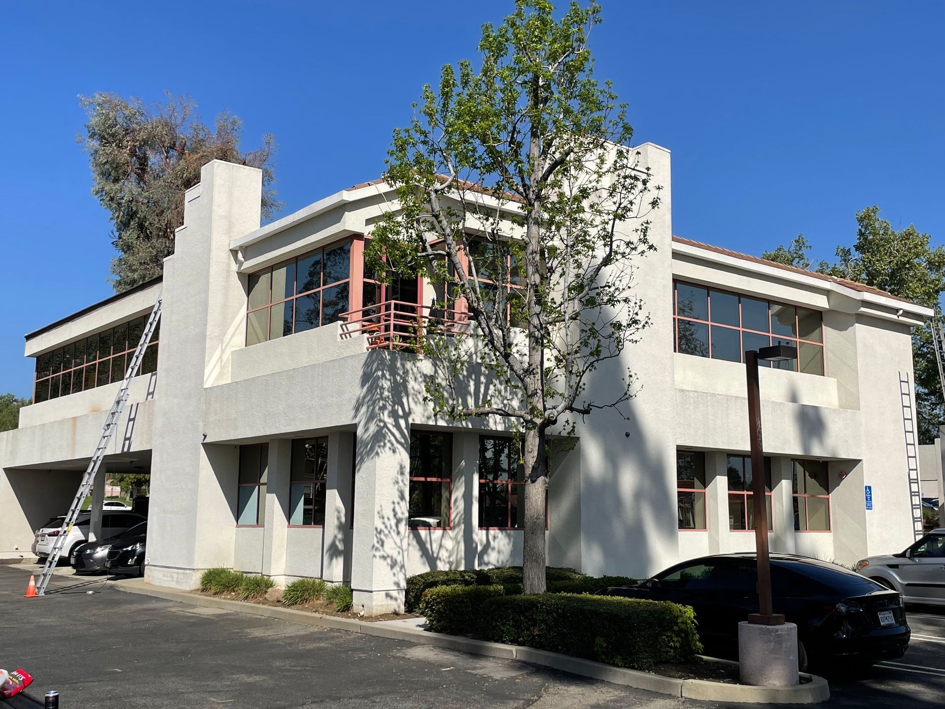Two-story white building with dark tinted windows. Cars in front, ladder on side, tree in front. Blue sky.