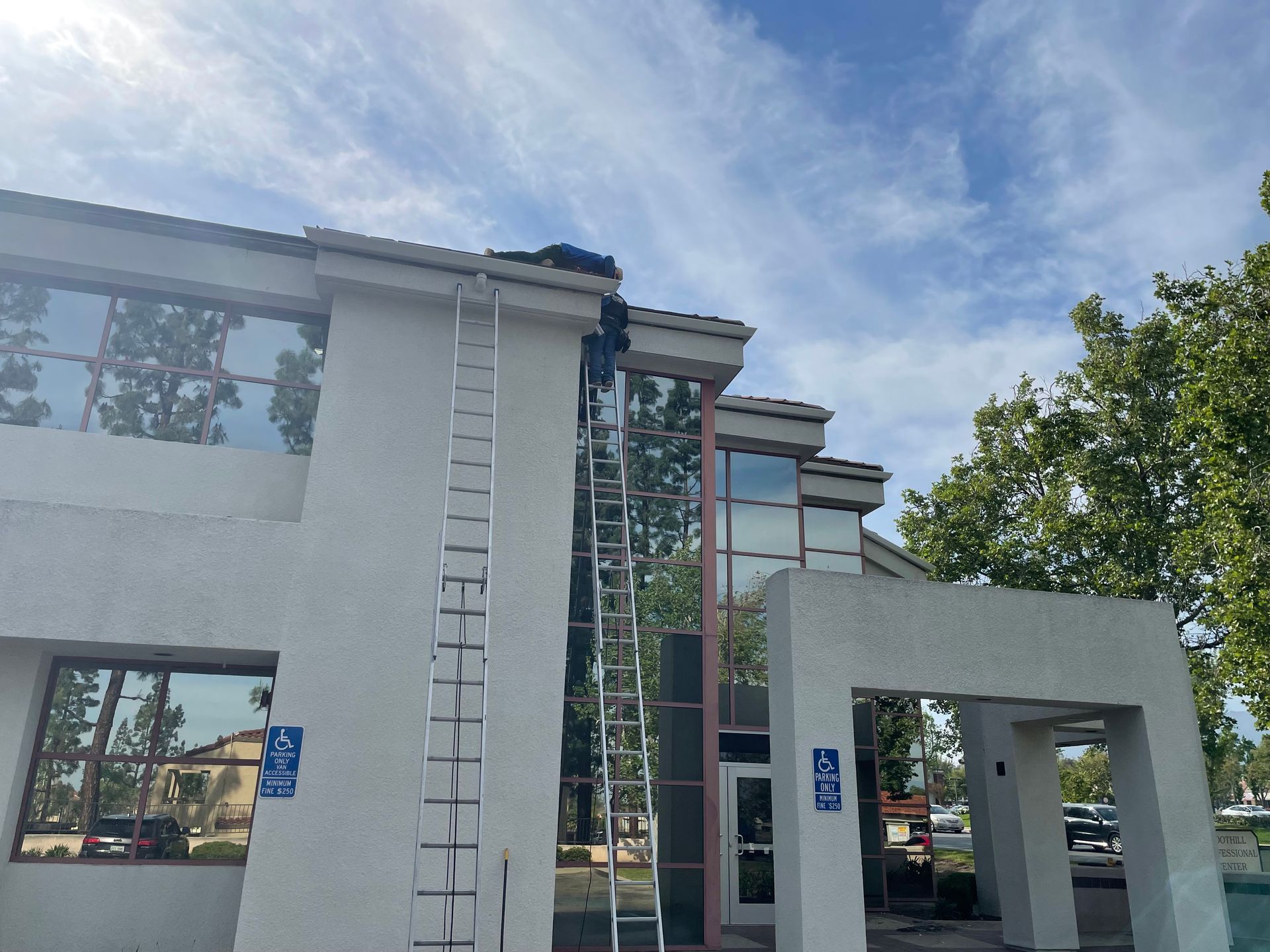 Person on a tall ladder working on a building roof. Building has windows, a ramp symbol, and trees.