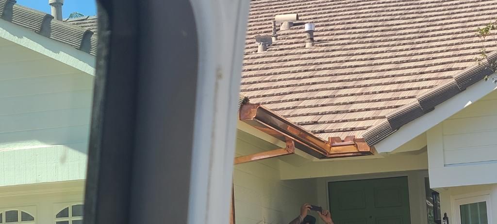 View of a house with brown roof tiles and a brown gutter, visible from a window.