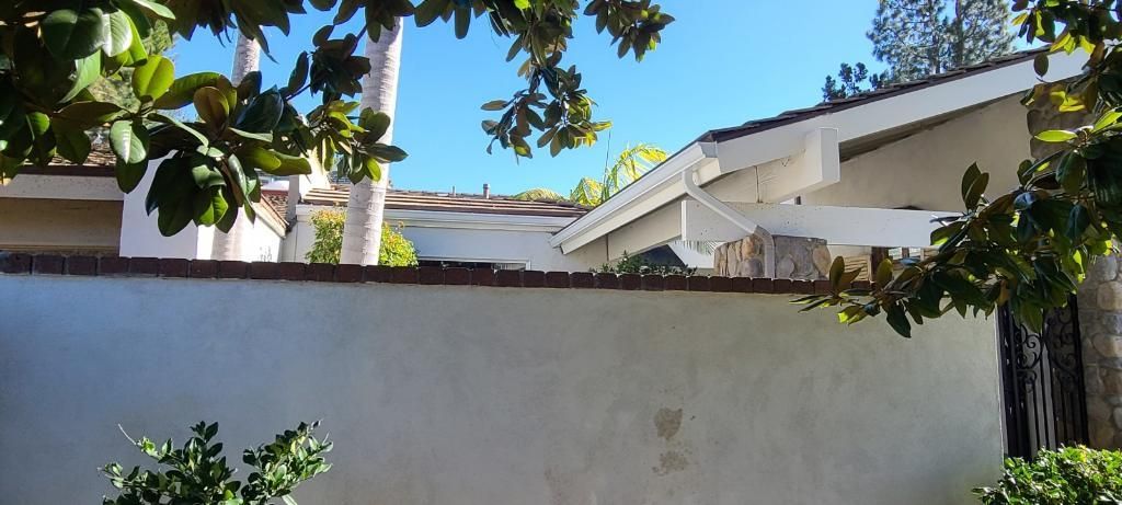 A white wall with a brick top, framing part of a house and trees with a clear blue sky above.
