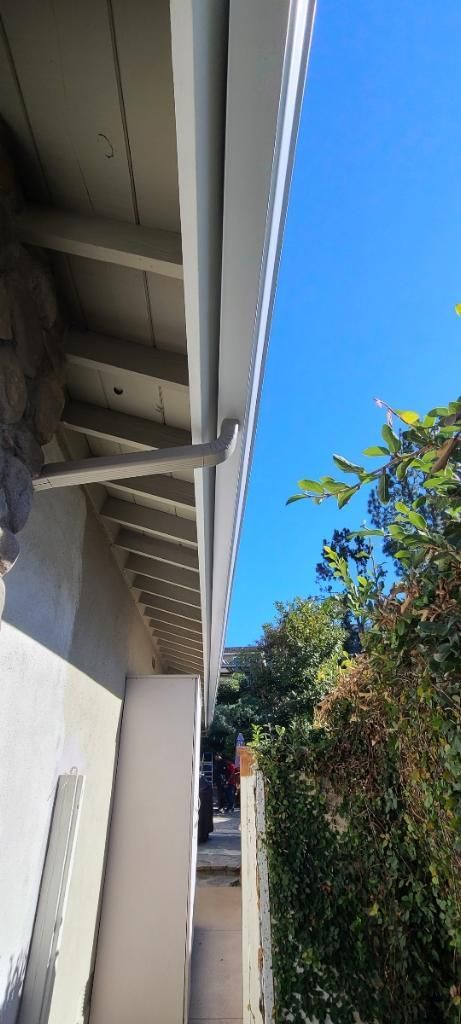 White roof gutter next to a building with a stone wall and green bush with blue sky.