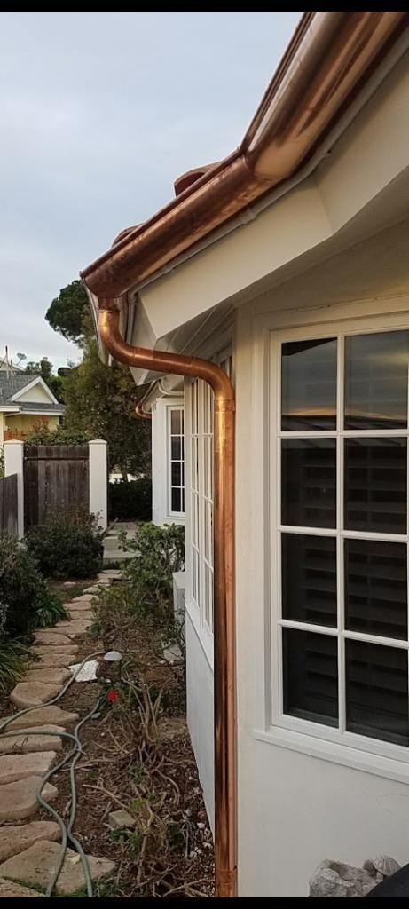 Copper gutters on a white house with a stone walkway, shrubs, and a fence in the background.