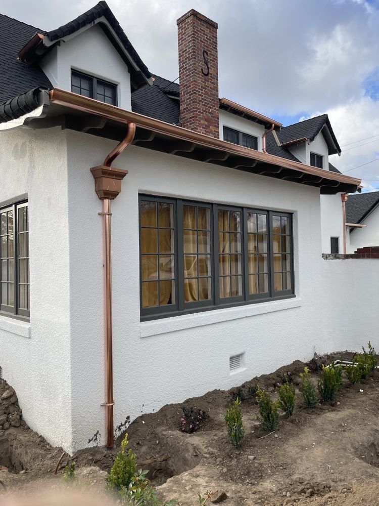 Copper gutters and downspout on a white stucco house, with a dark roof and windows.