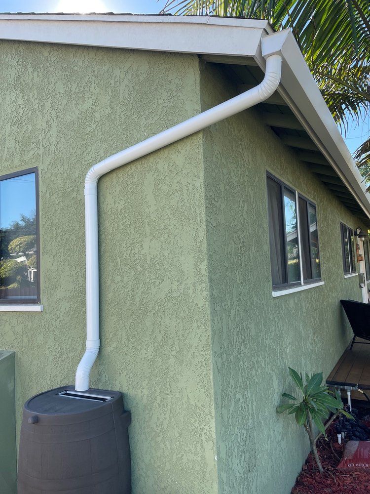 A white gutter system directs rainwater into a dark rain barrel next to a green stucco house.