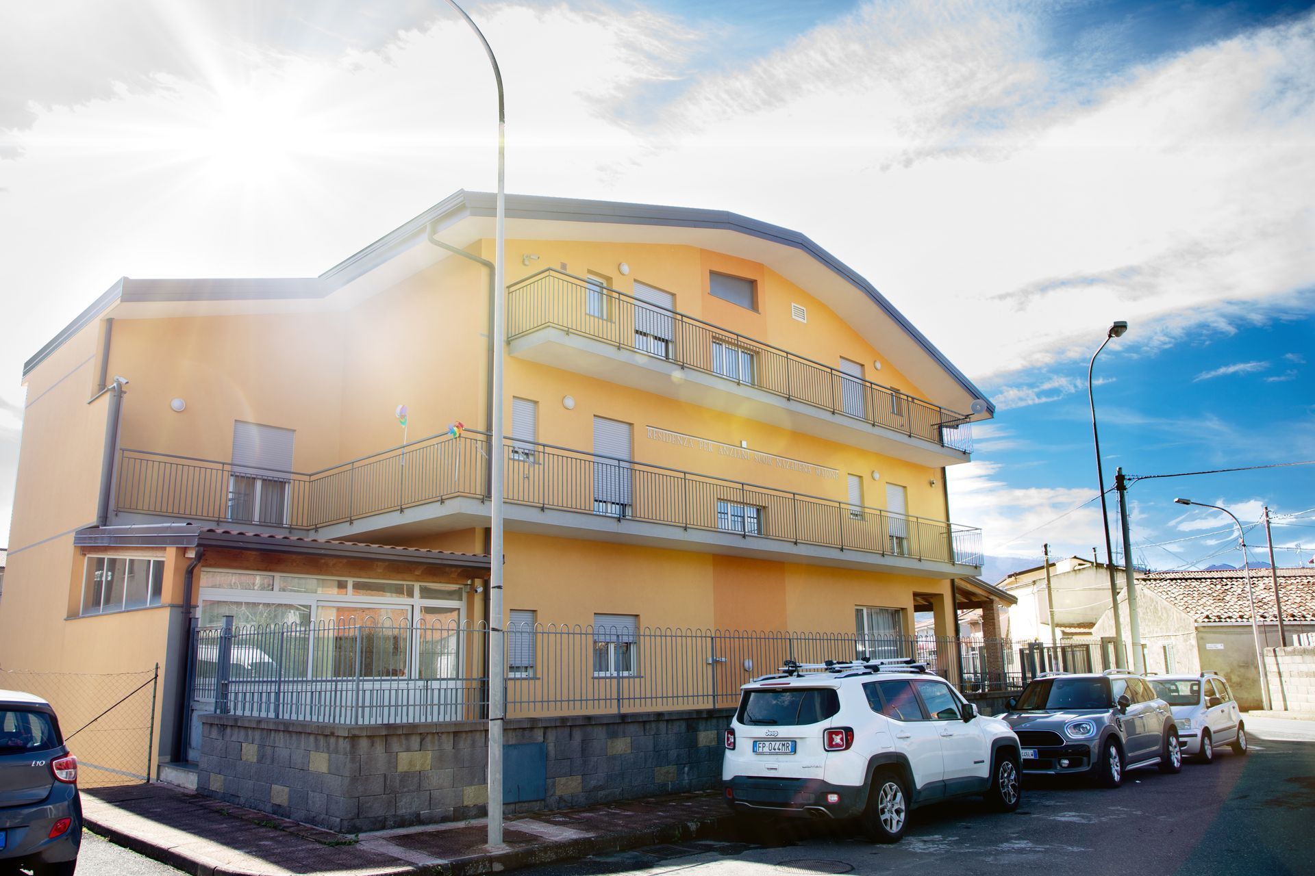 Edificio giallo con balconi, auto parcheggiate su una strada sotto un cielo parzialmente nuvoloso.