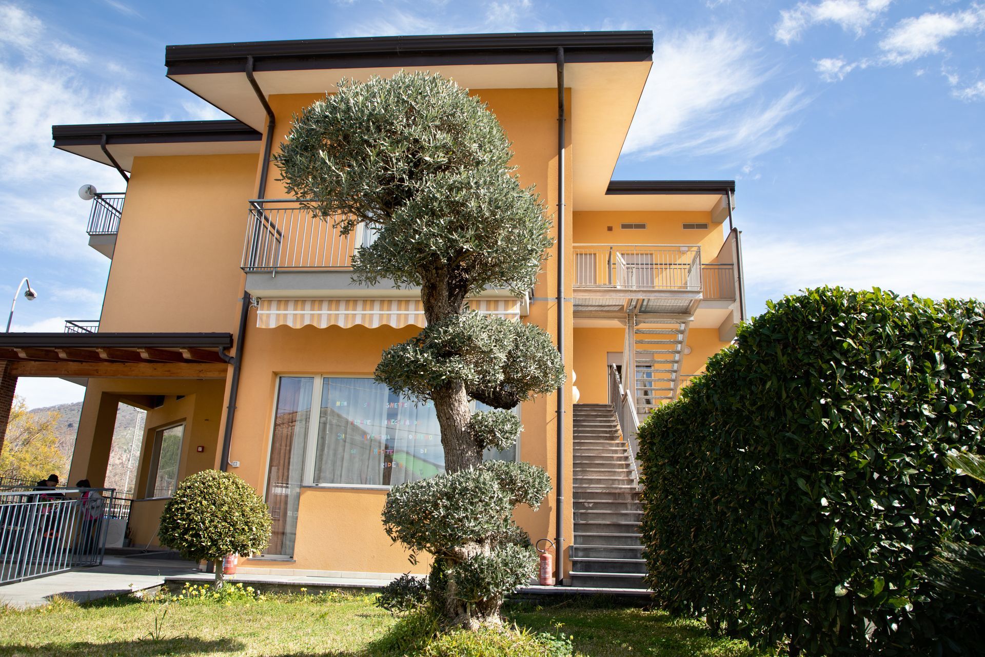 Edificio giallo a due piani con balconi, scala esterna, alberi ben curati e siepi che si stagliano contro il cielo azzurro.