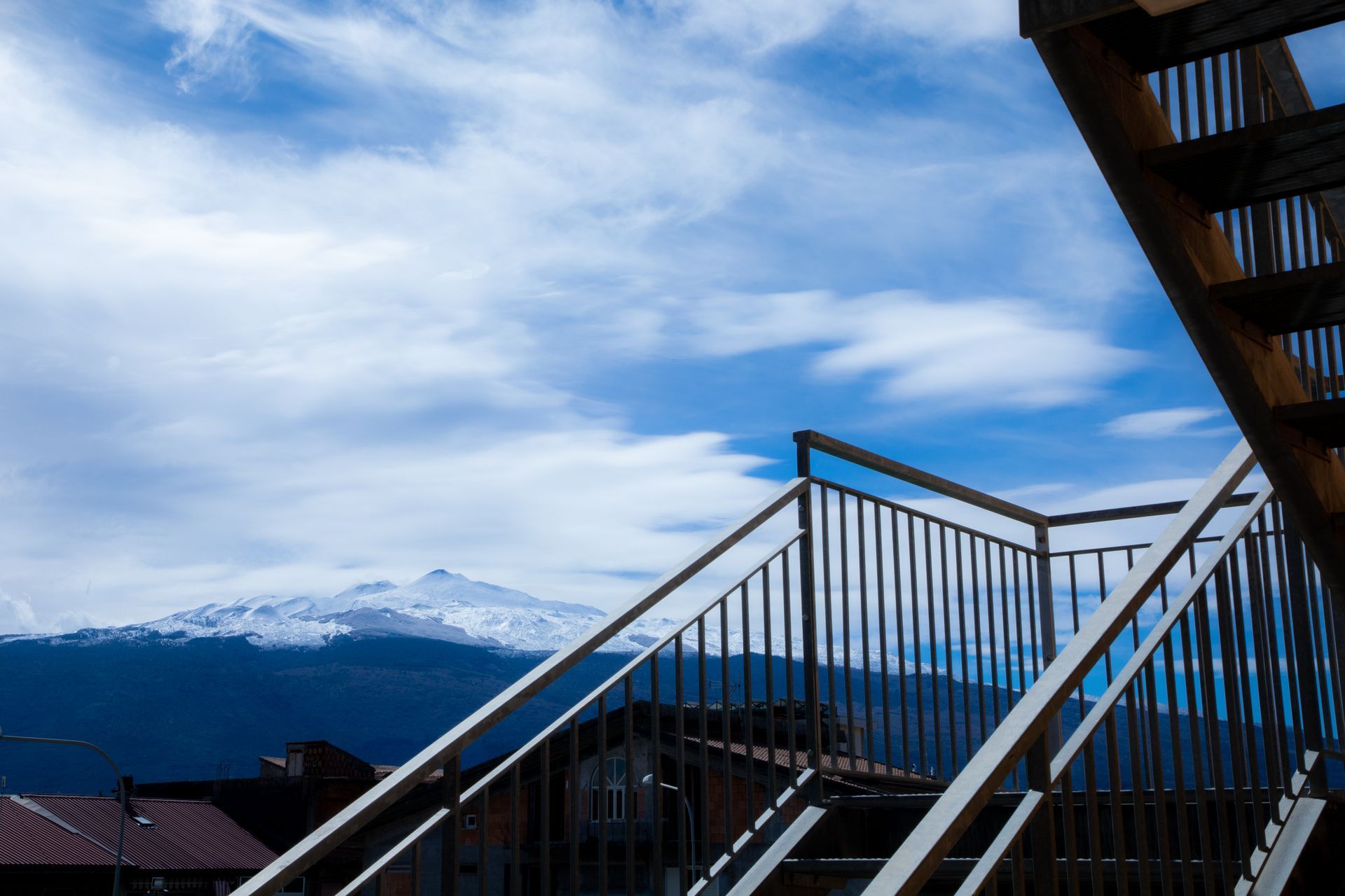 Scalinata che sale con vista sulle montagne e cielo nuvoloso. Ringhiere scure di fronte alle cime innevate.