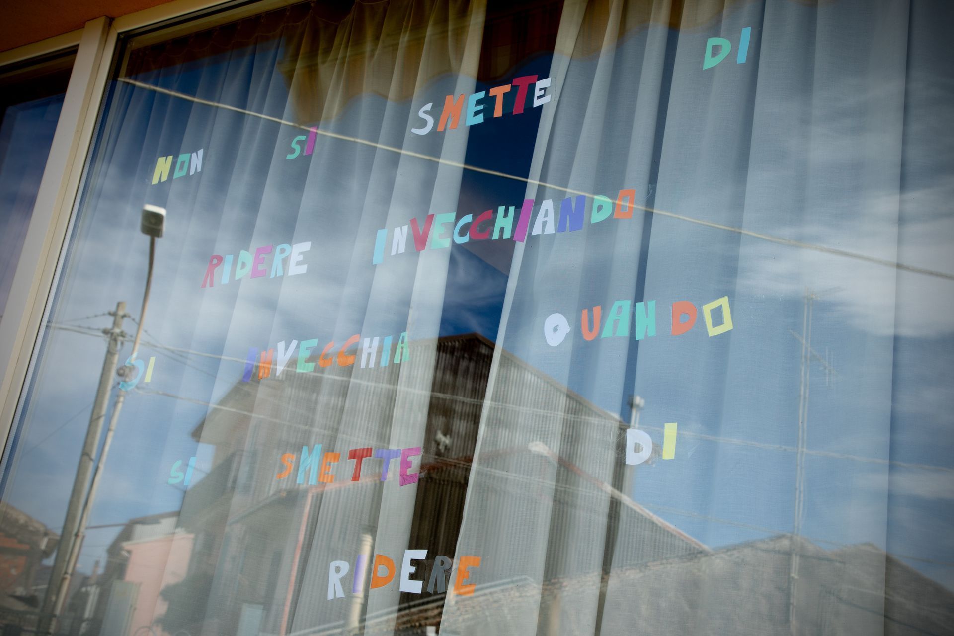 Finestra con lettere colorate che compongono una frase in italiano, in cui si riflettono edifici e cielo.