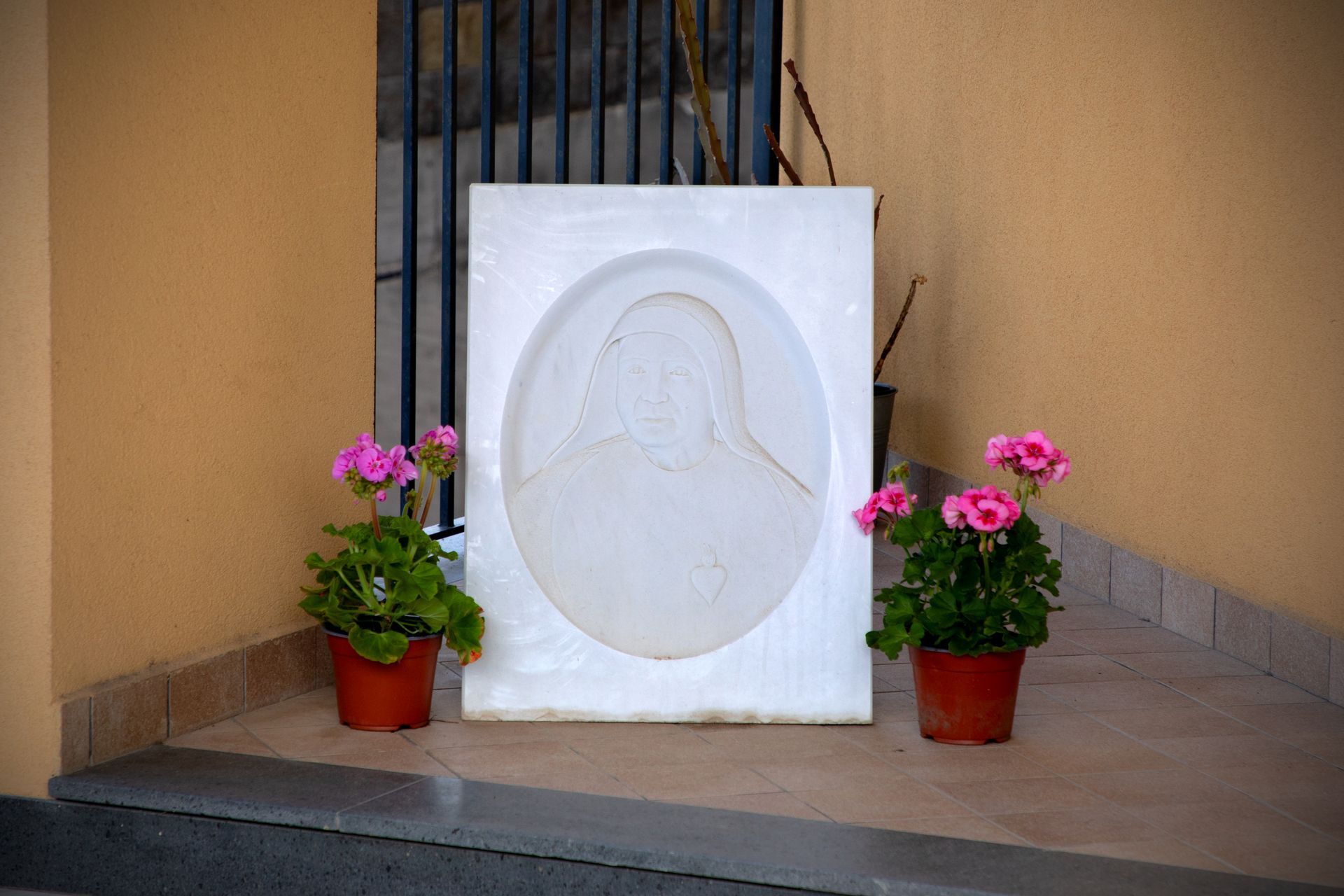 Rilievo in marmo raffigurante una figura velata, affiancata da gerani rosa in vaso.