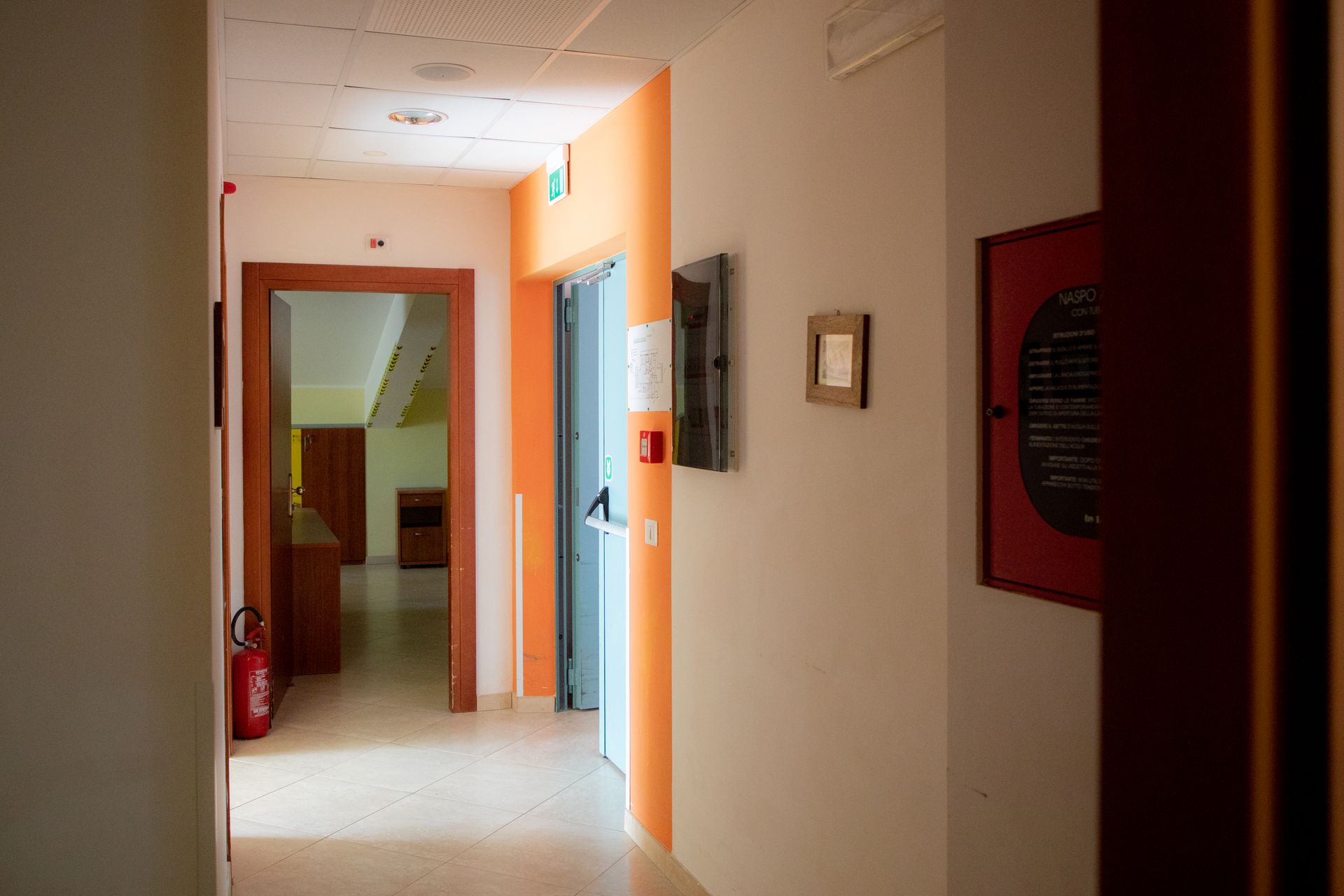 Corridoio con parete decorativa arancione, ascensore e porte. Estintore sulla sinistra.