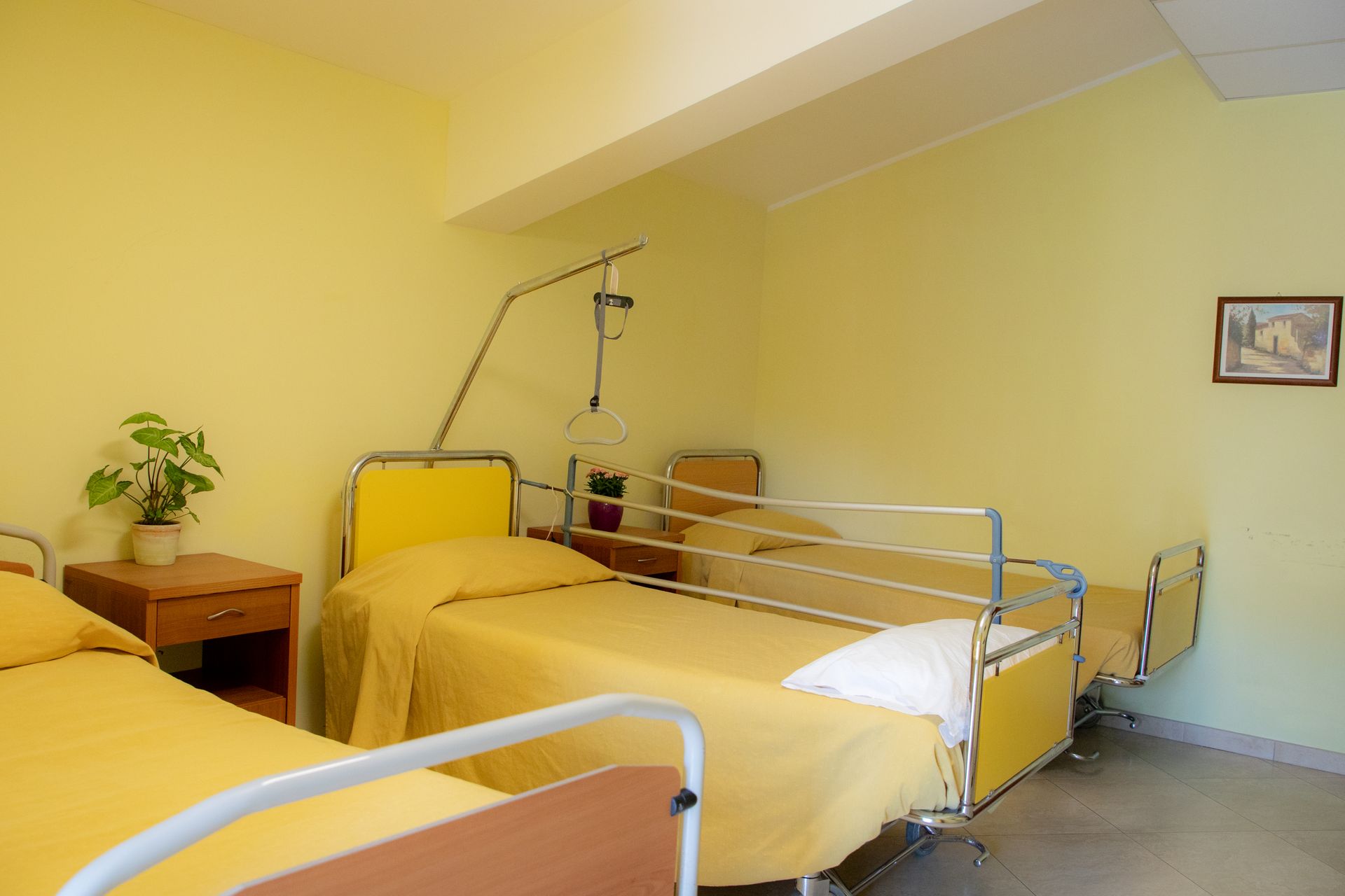 Stanza d'ospedale con tre letti gialli. Pareti gialle, un comodino e un piccolo quadro incorniciato.