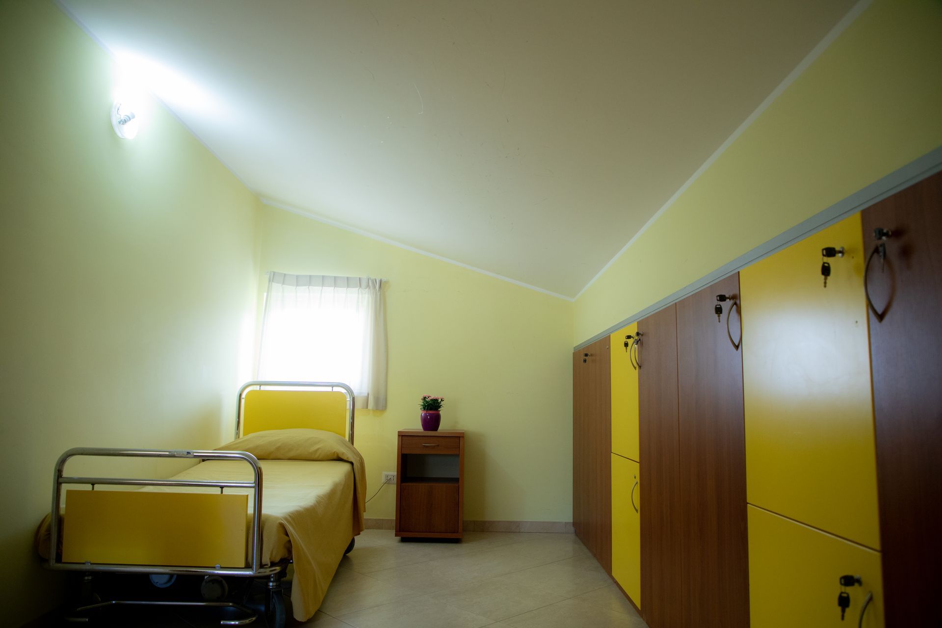 Una piccola stanza nei toni del giallo con un letto, degli armadietti, una finestra e un comodino.