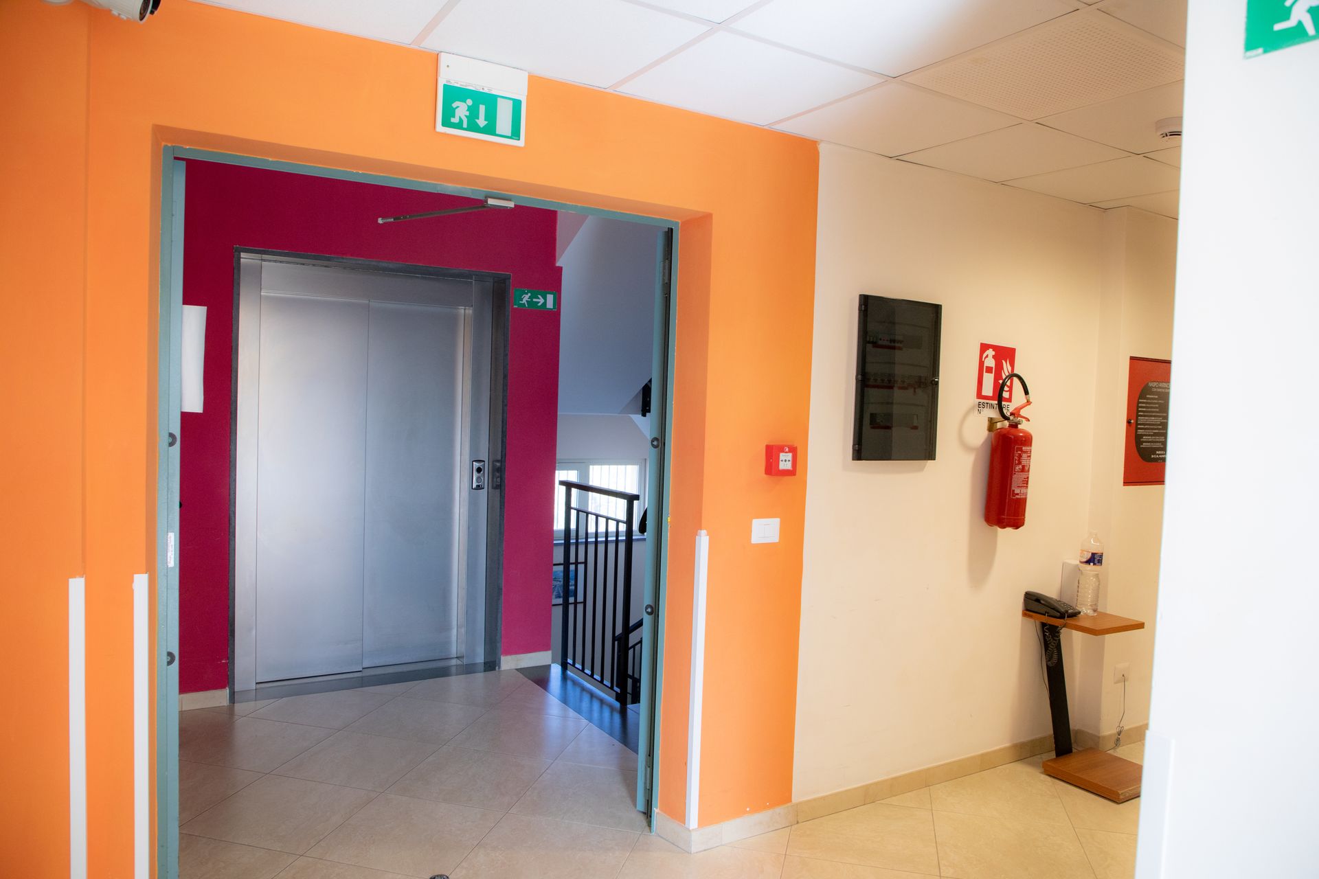 Corridoio che conduce a una porta aperta con una porta grigia, un estintore e segnali di uscita di emergenza.