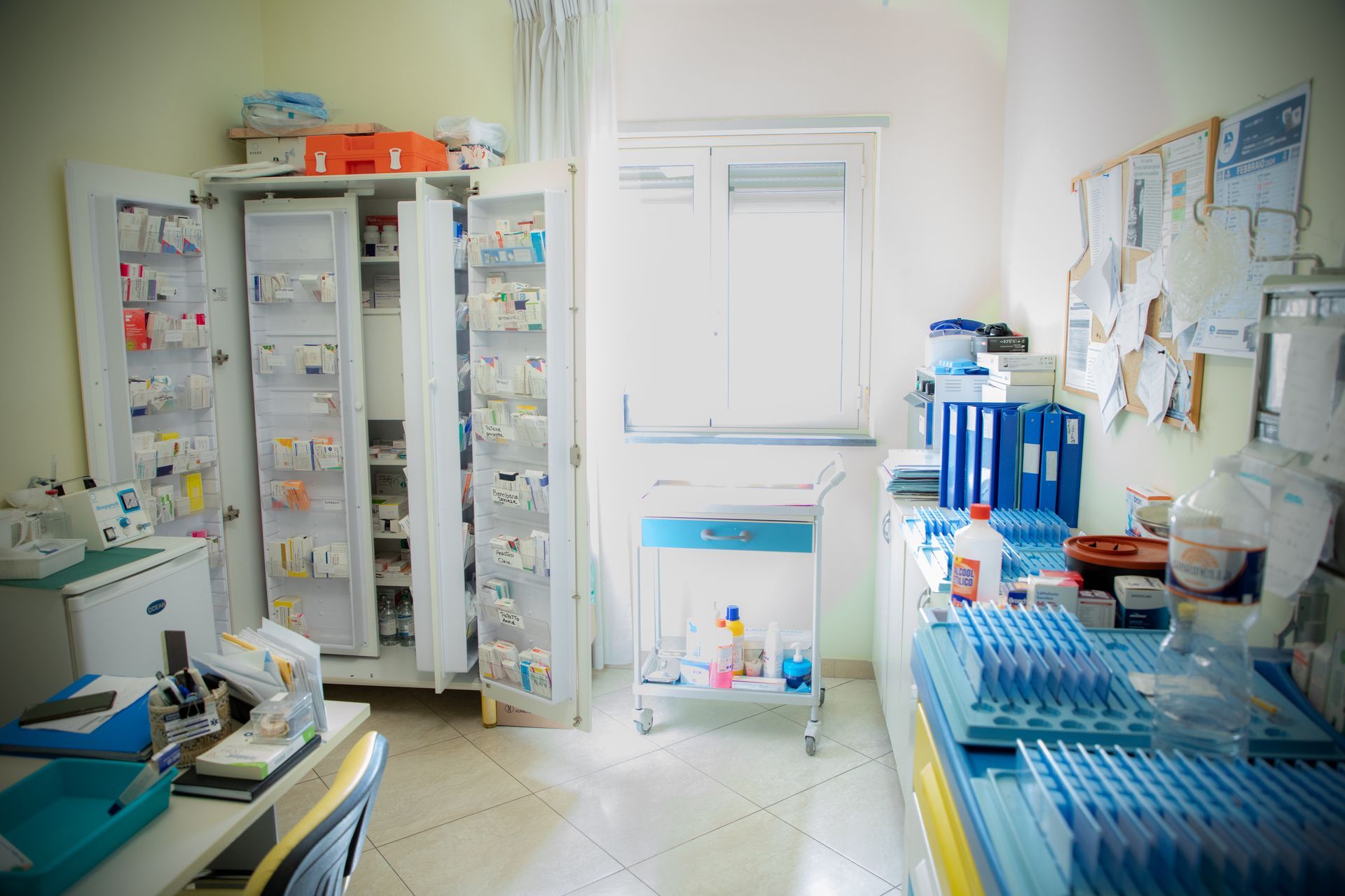 Un ripostiglio per forniture mediche disordinato, con armadietti, un carrello con ruote e una finestra.