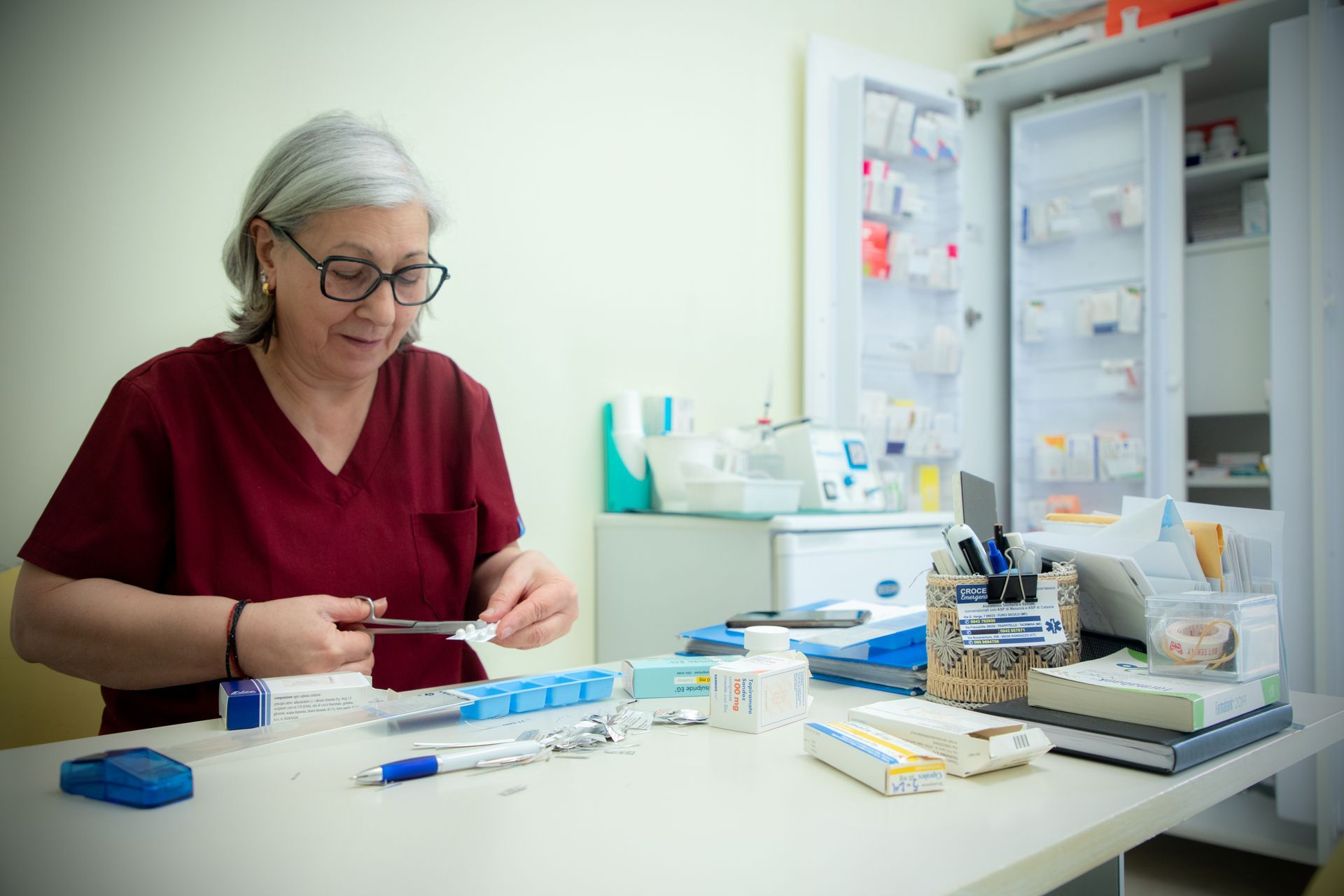 Una donna in camice bordeaux prepara i farmaci alla scrivania, sullo sfondo un armadietto dei medicinali aperto.