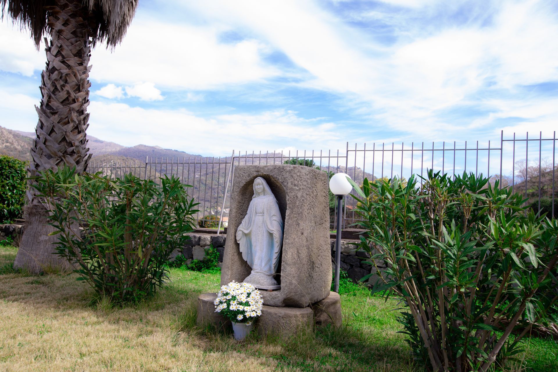 Statua della Vergine Maria in una nicchia di pietra, con montagne sullo sfondo sotto un cielo nuvoloso.