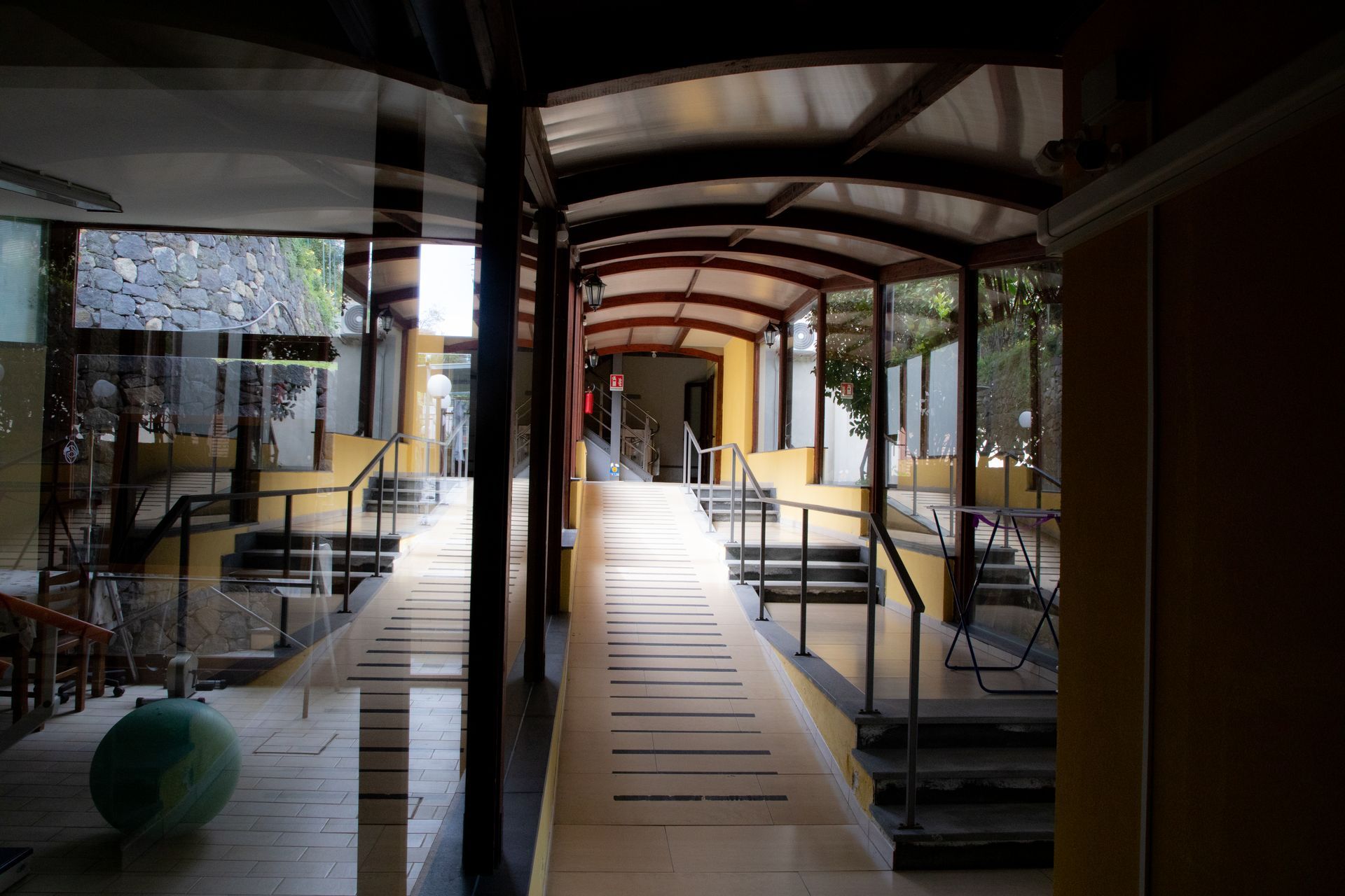 Passaggio coperto con rampa, scale e parete in vetro. Toni gialli e marroni, luce intensa all'estremità.