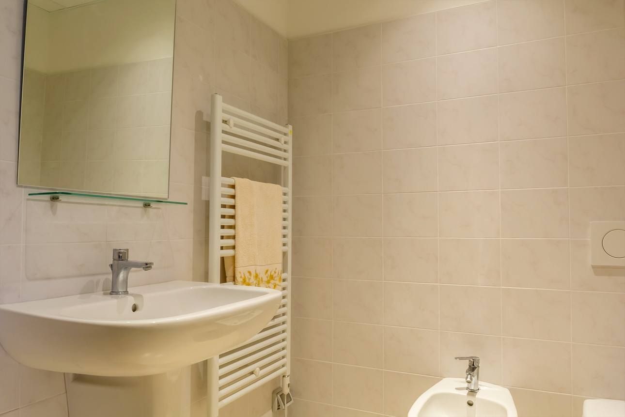 Bagno con lavandino bianco, specchio, portasciugamani e bidet; pareti piastrellate.