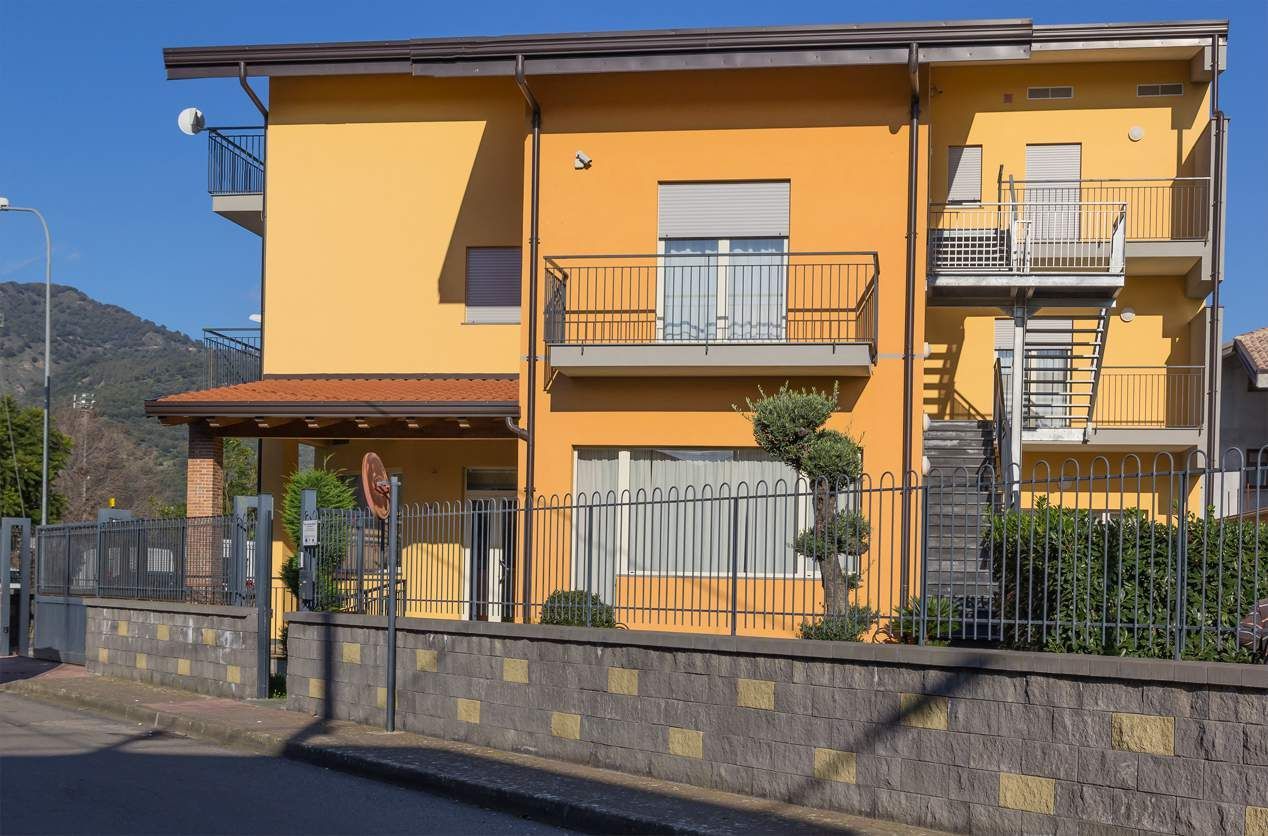 Edificio giallo a due piani con balconi, situato dietro una recinzione e un muretto basso.