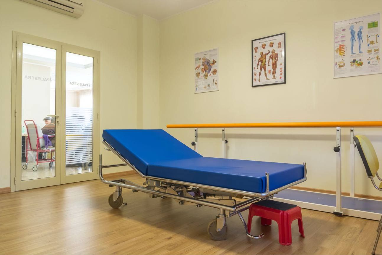 Sala visita medica: letto blu per pazienti, cartelle cliniche sul muro, porta, sgabello rosso, corrimano giallo.