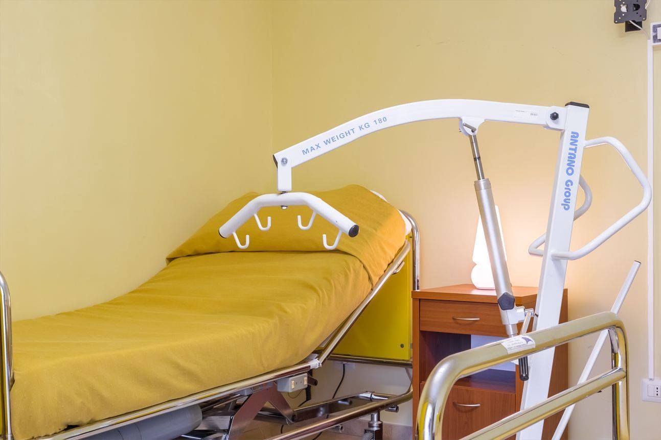 Letto d'ospedale con sollevatore per pazienti. Lenzuola e pareti giallo chiaro.