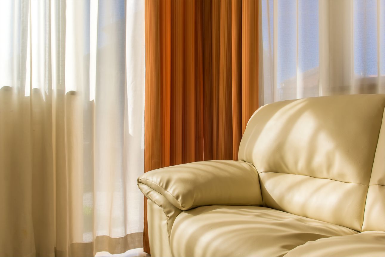 Divano in pelle color crema accanto alla finestra con tende bianche e arancioni trasparenti.
