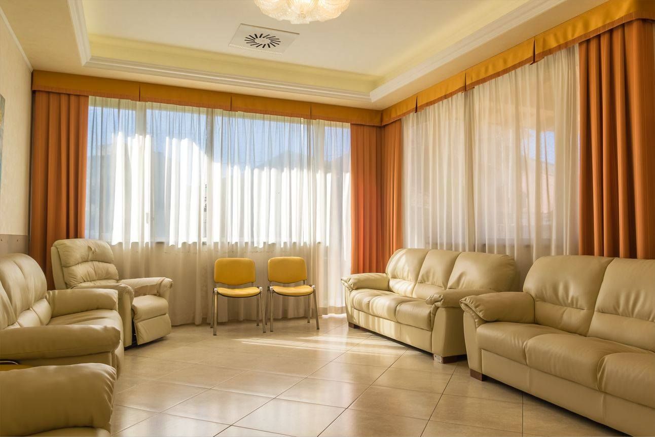 Sala d'attesa luminosa con divani color crema, tende arancioni e luce del sole che filtra attraverso tende.