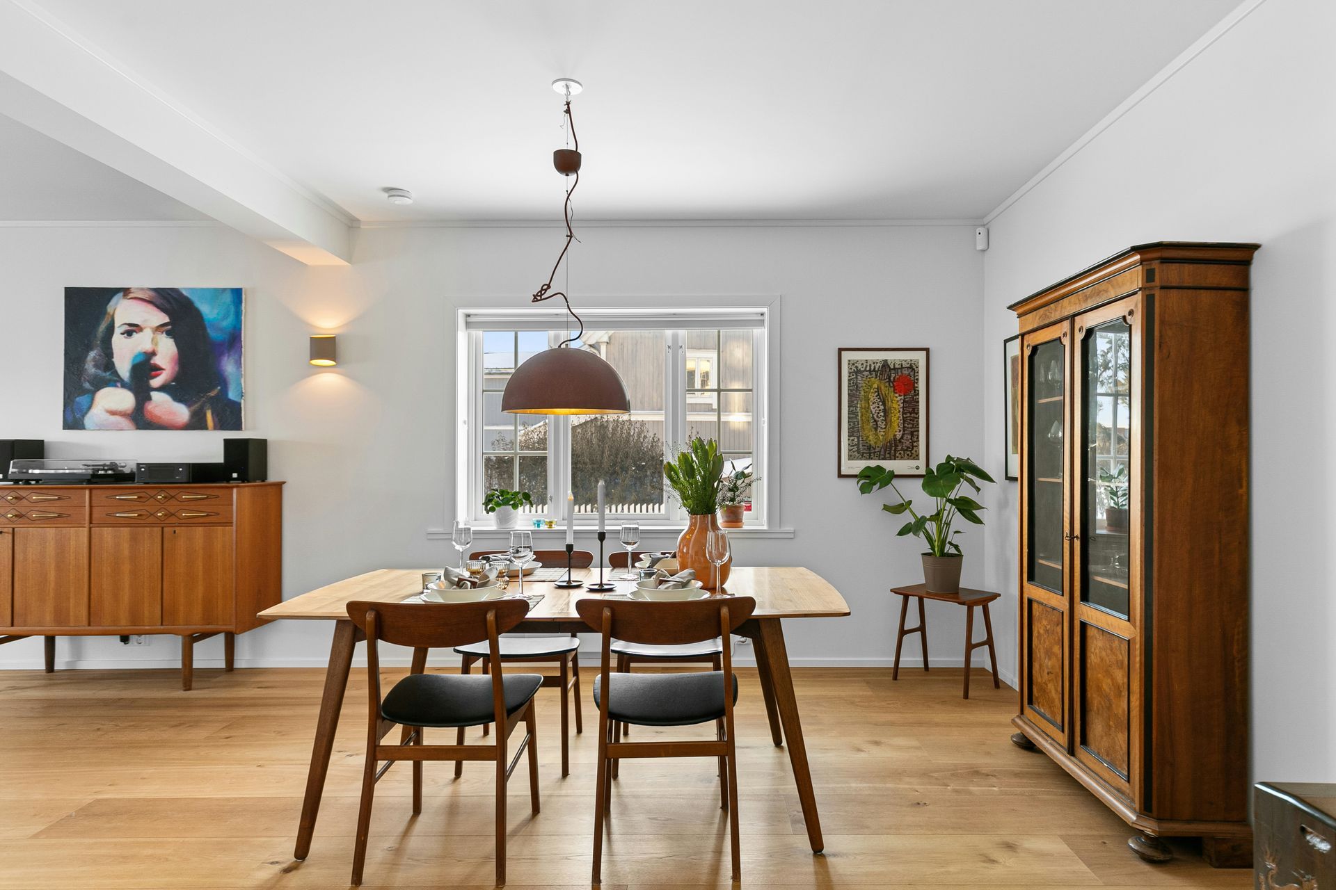 Bright dining room with wooden table and chairs, sideboard, cabinet, plants, and a large window.