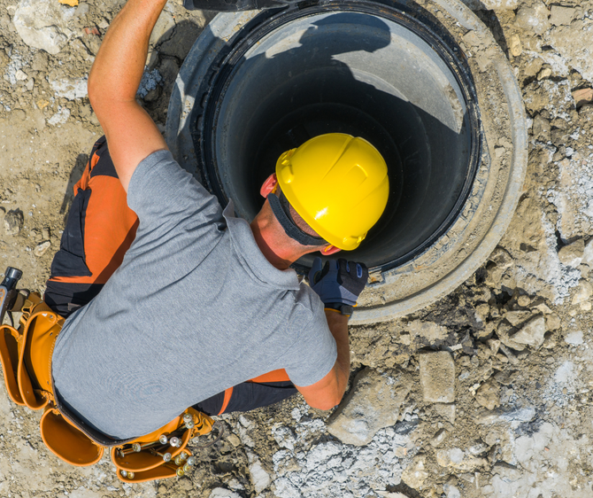 Construction worker in yellow hard hat examining a large pipe in a concrete opening.