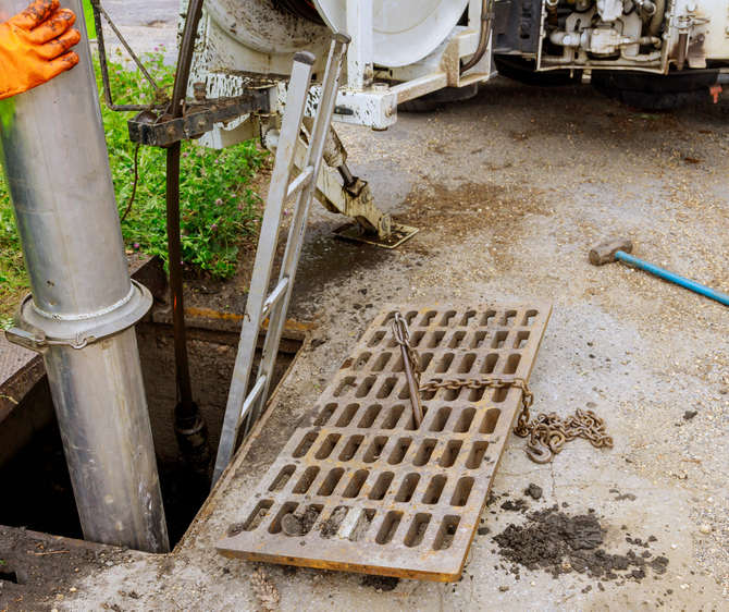 Sewer maintenance: worker in orange glove lowering equipment into an open manhole. Ladder and grate nearby.