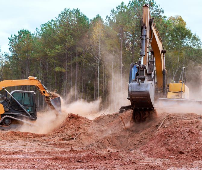 Two excavators digging earth in a construction site, creating dust clouds; trees in the background.