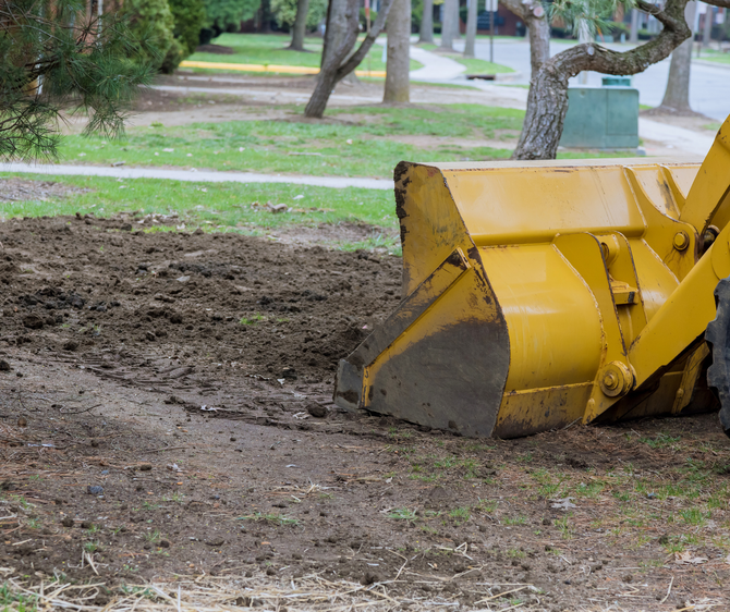 Yellow front-end loader on dirt in a grassy area, clearing soil near a sidewalk.